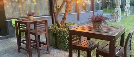 An outdoor patio area with wooden tables and chairs, a potted plant, and a view of a restaurant through a glass window.