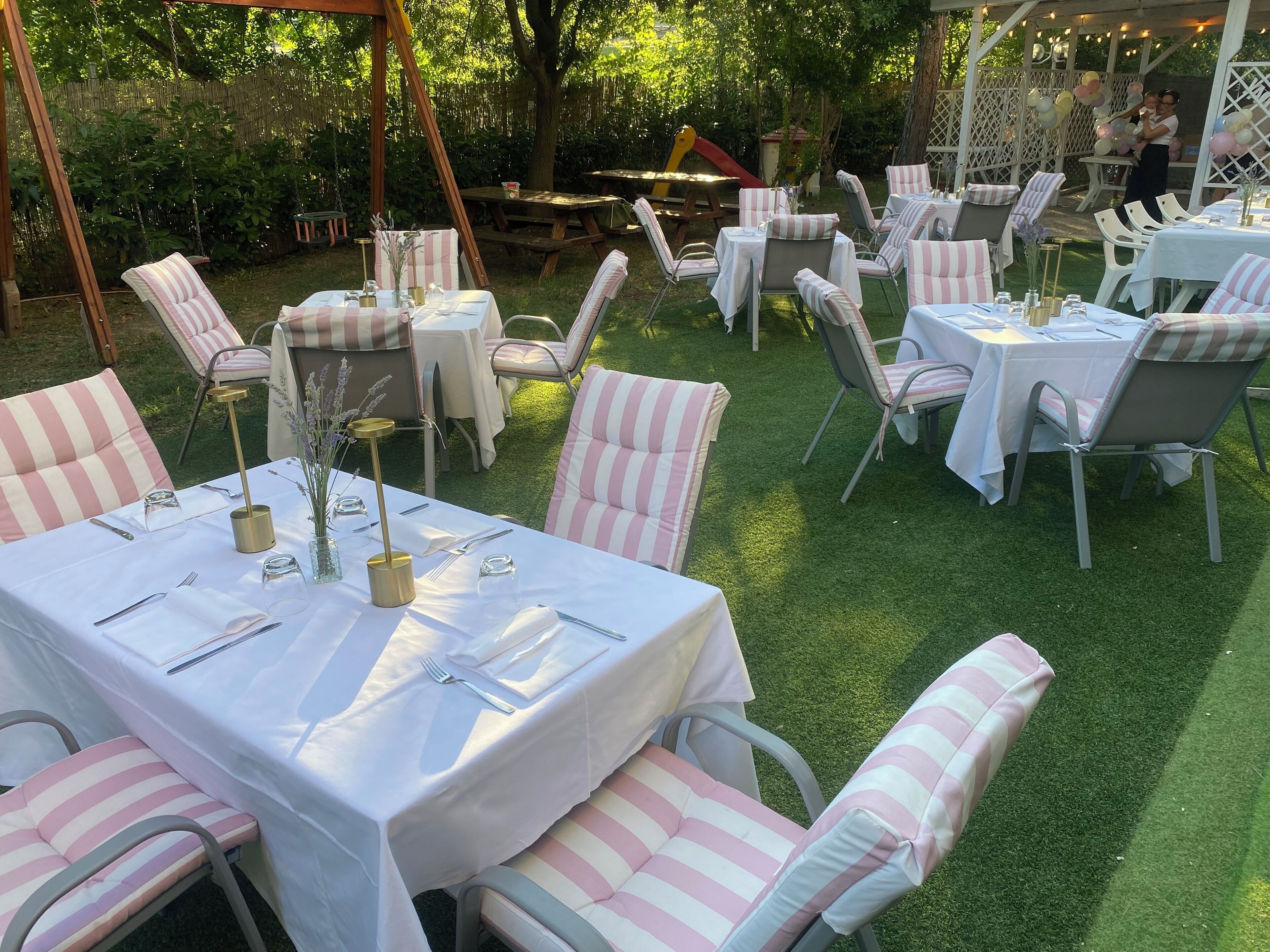 An outdoor dining area with tables and chairs, surrounded by greenery and a wooden structure.