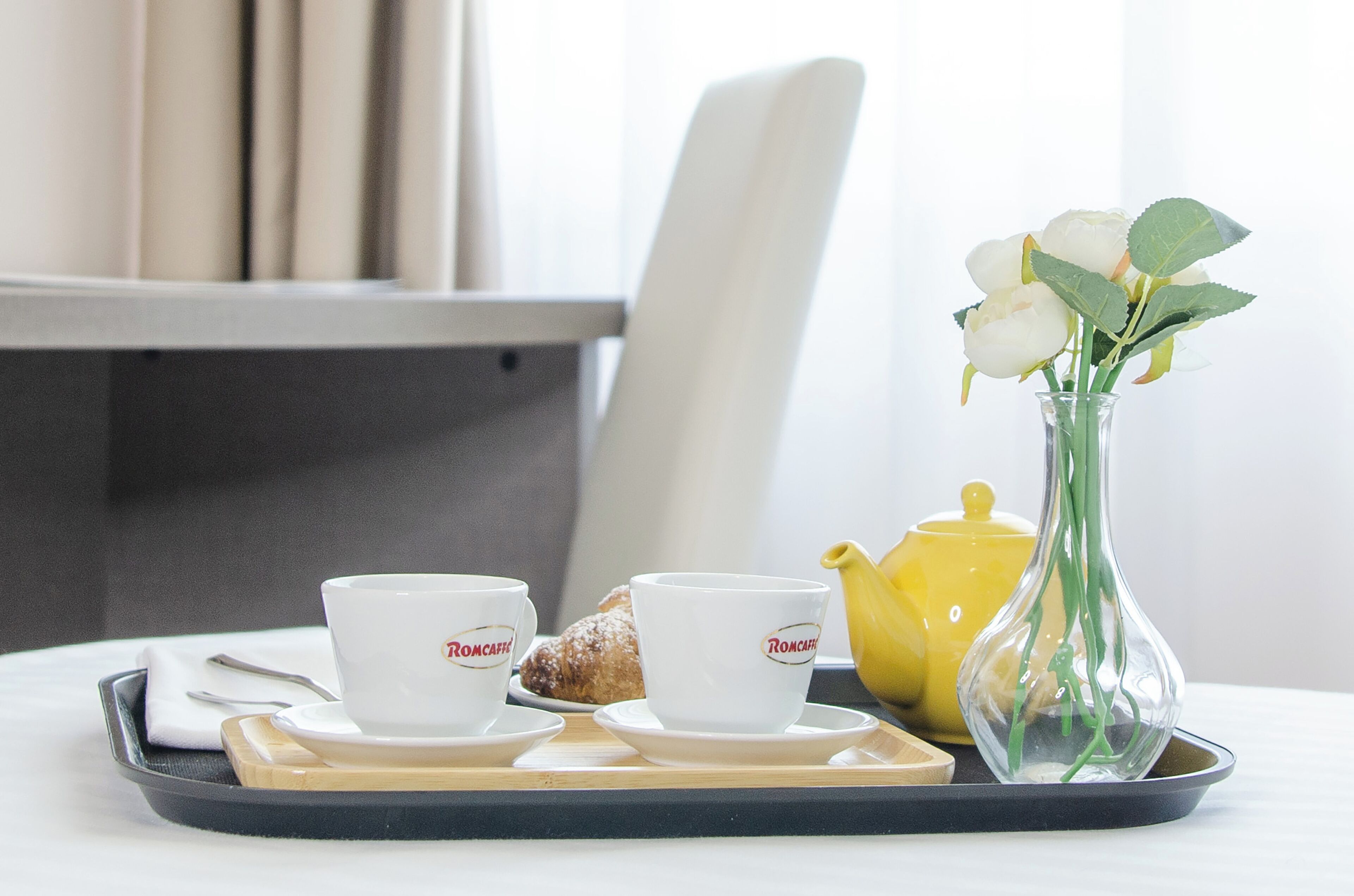 A tray with two white cups, a yellow teapot, a croissant, and a vase with white flowers.