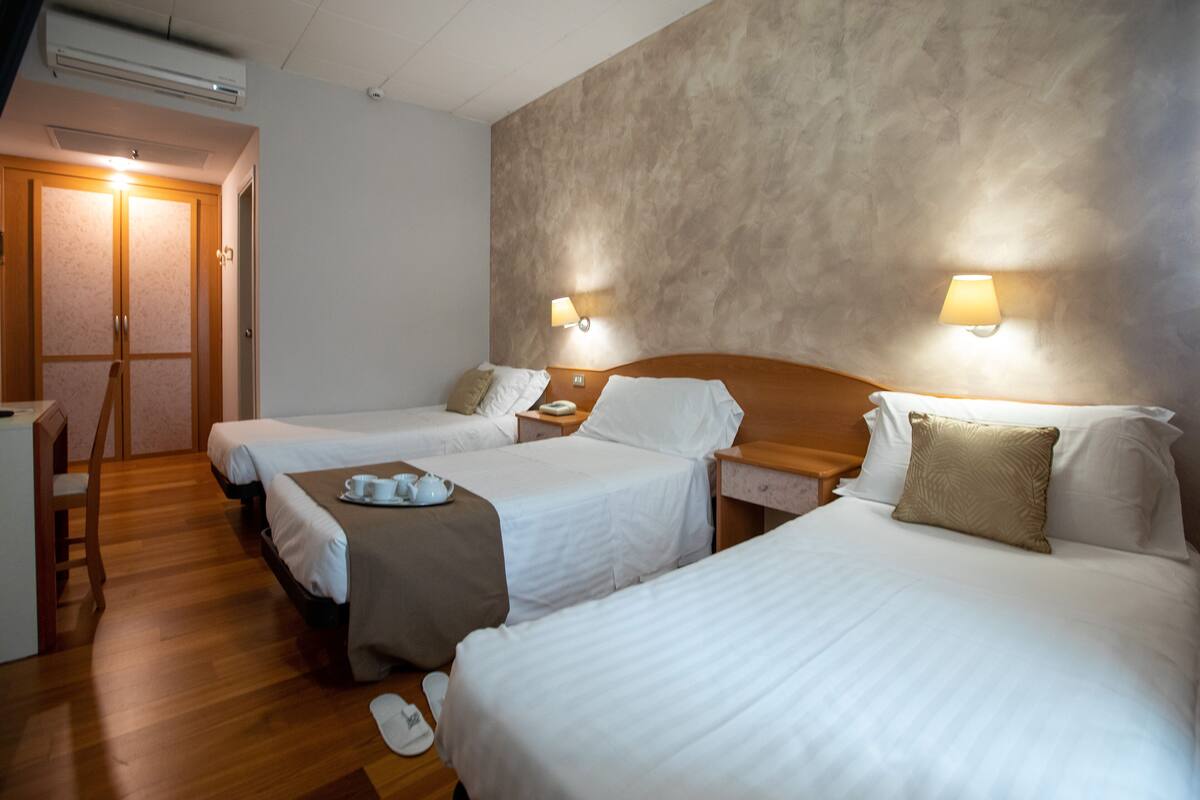 A hotel room with two beds, wooden flooring, a desk with a chair, and a wall-mounted lamp.