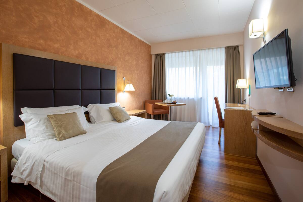 A hotel room with a large bed, a desk, a chair, a TV, and a window with curtains.