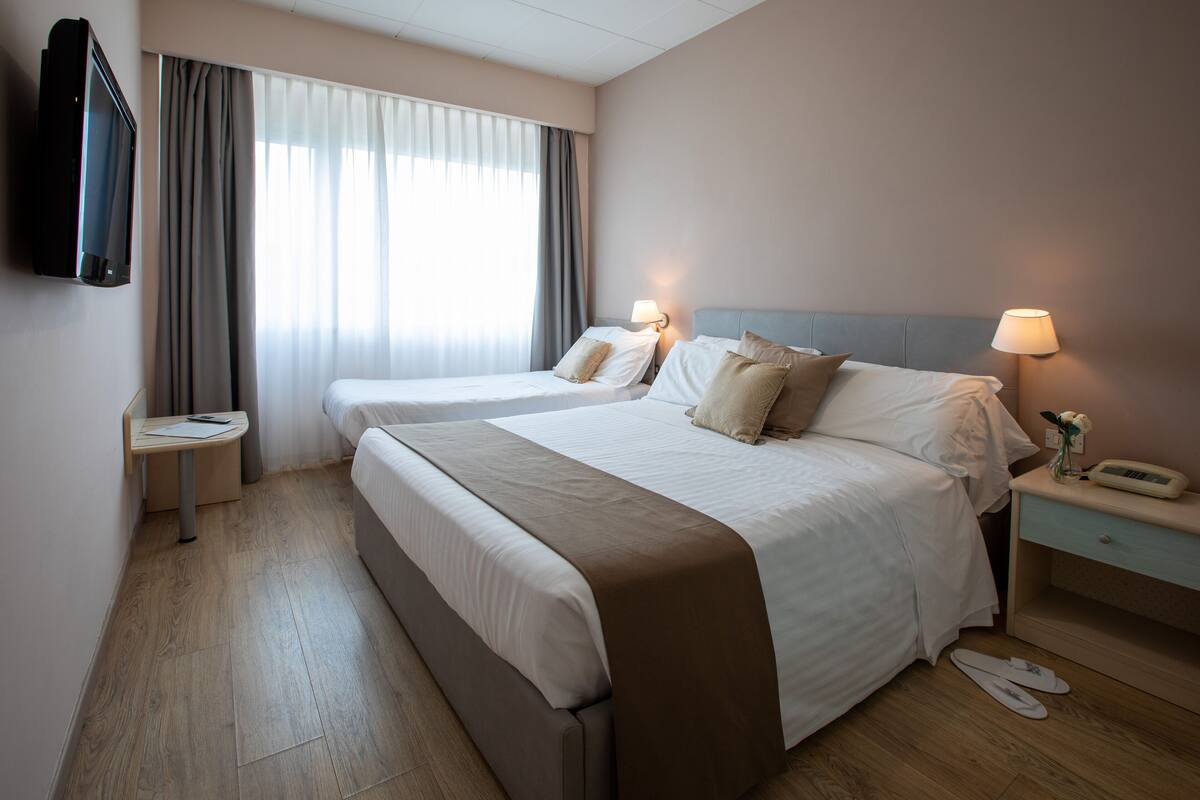 A hotel room with two beds, a TV mounted on the wall, a bedside table with a lamp, and a small table with a remote control.