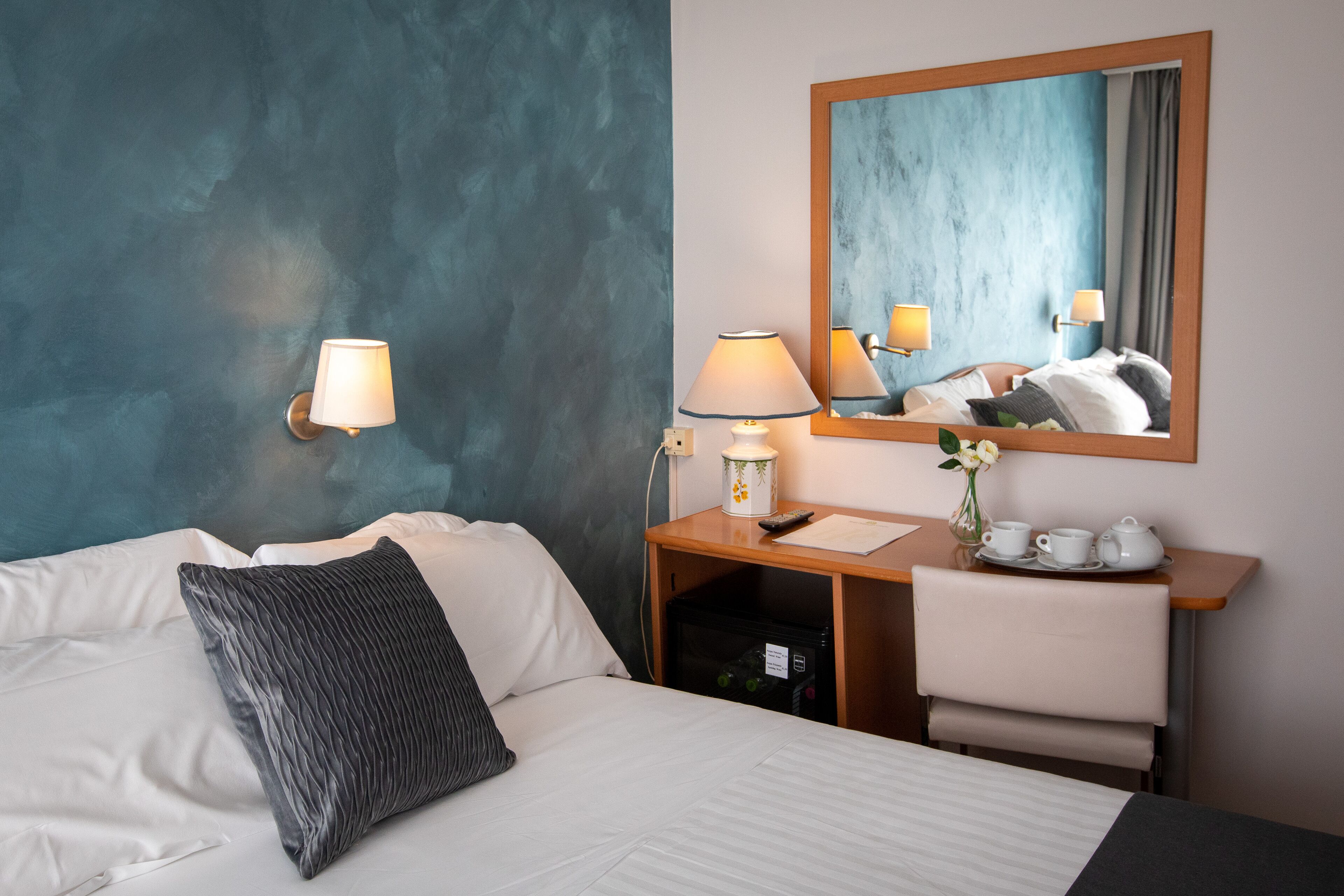 Habitación de hotel con cama, escritorio con lámpara, espejo y un jarrón con flores.