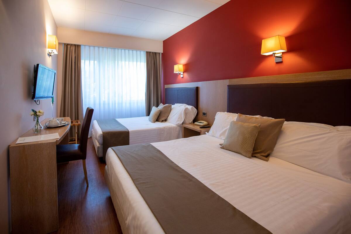 A hotel room with two beds, a desk, a chair, a TV, and a lamp.