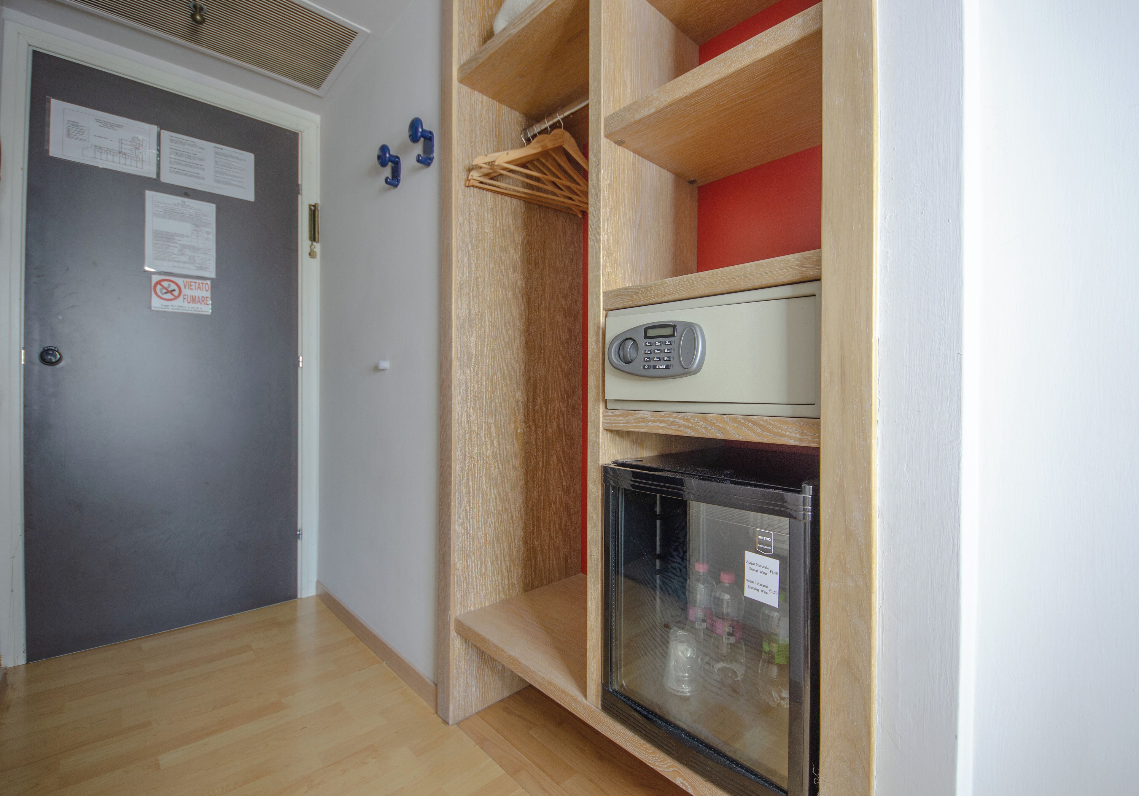 A compact room with a grey door, a small fridge, and a wooden shelf unit with a red item on top.