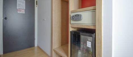A compact room with a grey door, a small fridge, and a wooden shelf unit with a red item on top.