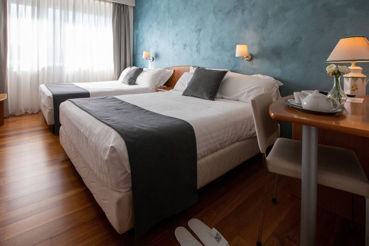 A hotel room with two beds, a wooden desk with a lamp, and a view of the city through large windows.