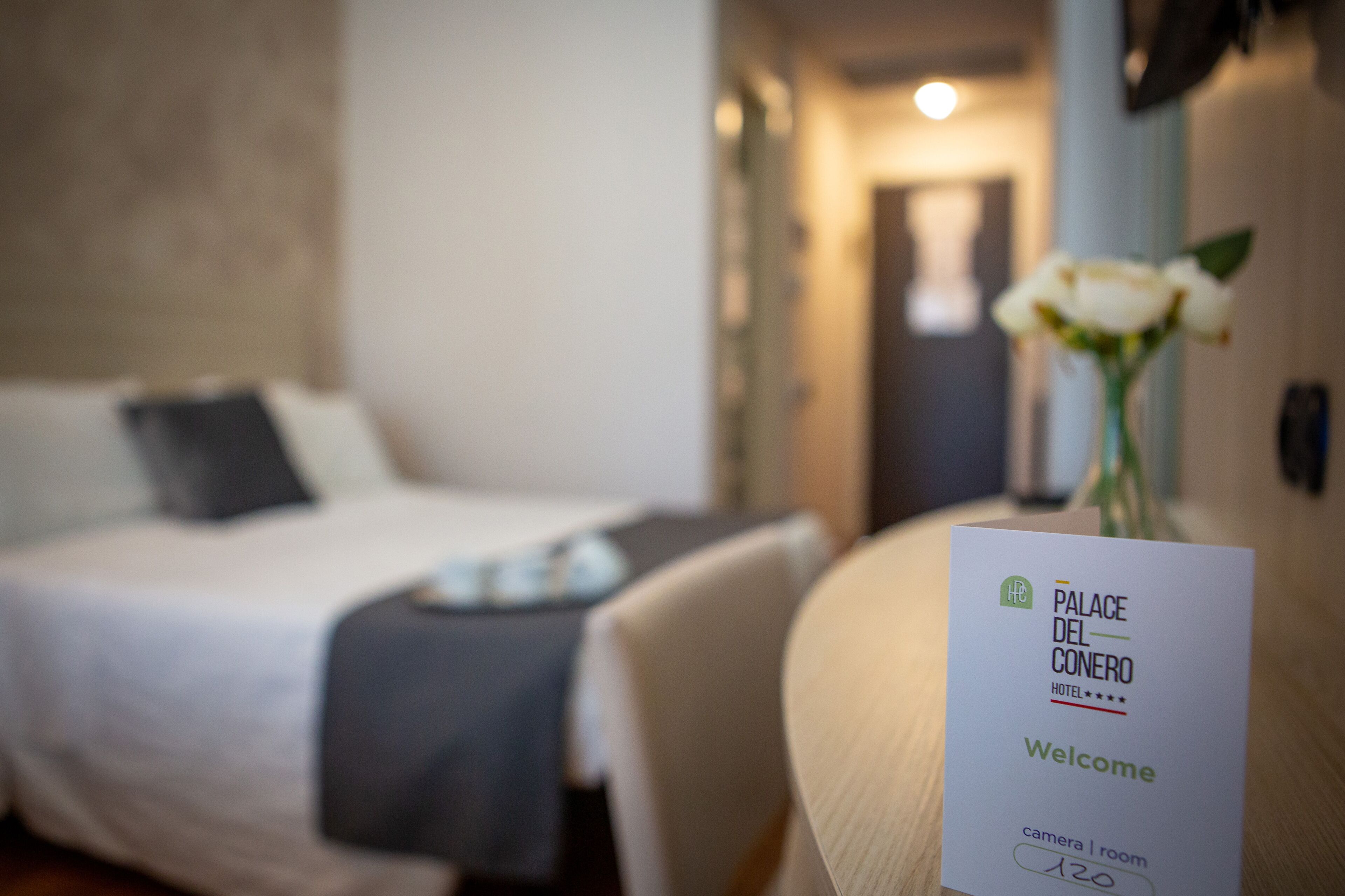 A hotel room with a bed, a bedside table, a vase with flowers, and a welcome sign.