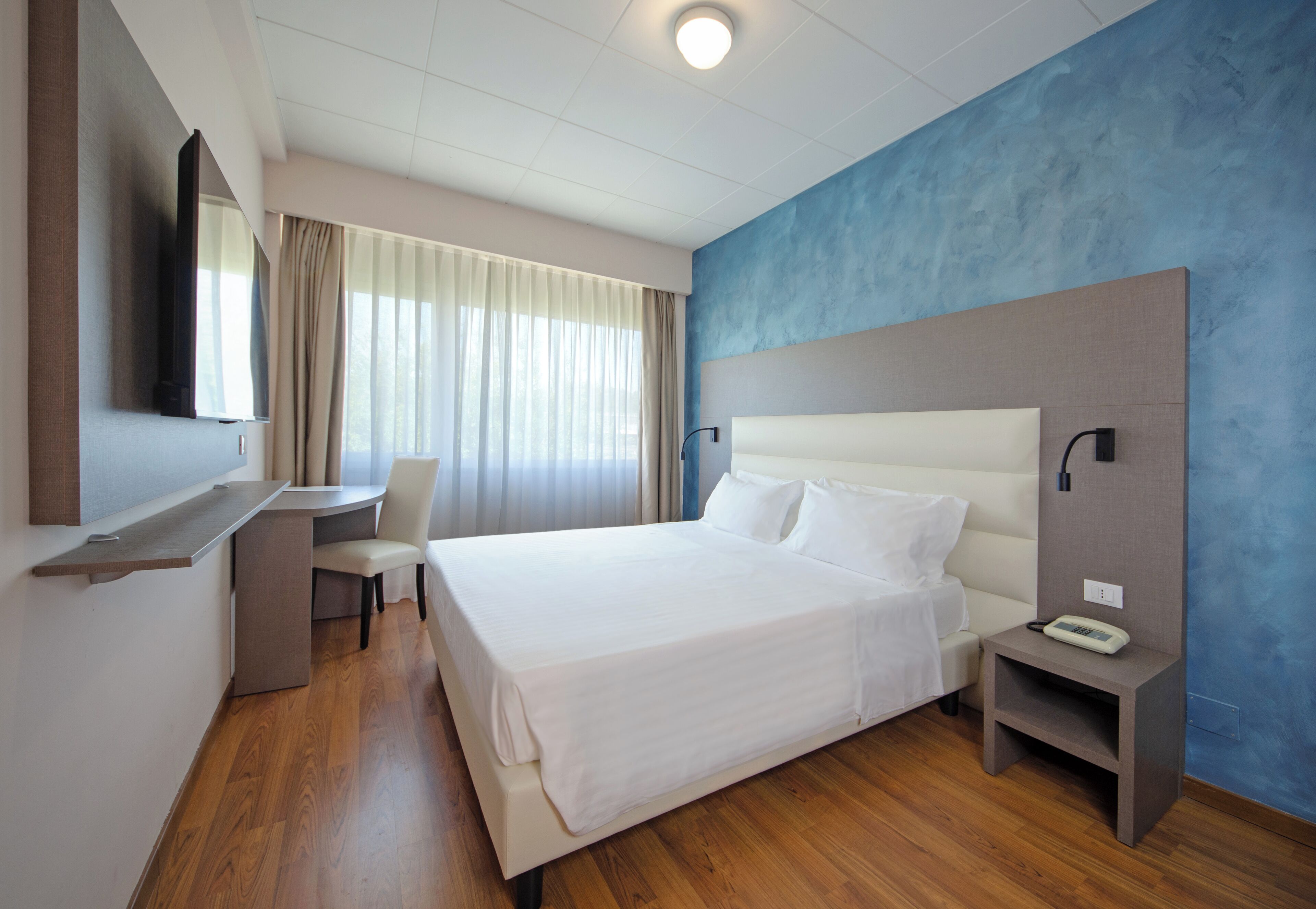Habitación de hotel con una cama grande, un escritorio, una silla, un televisor y un teléfono sobre una mesita de noche.