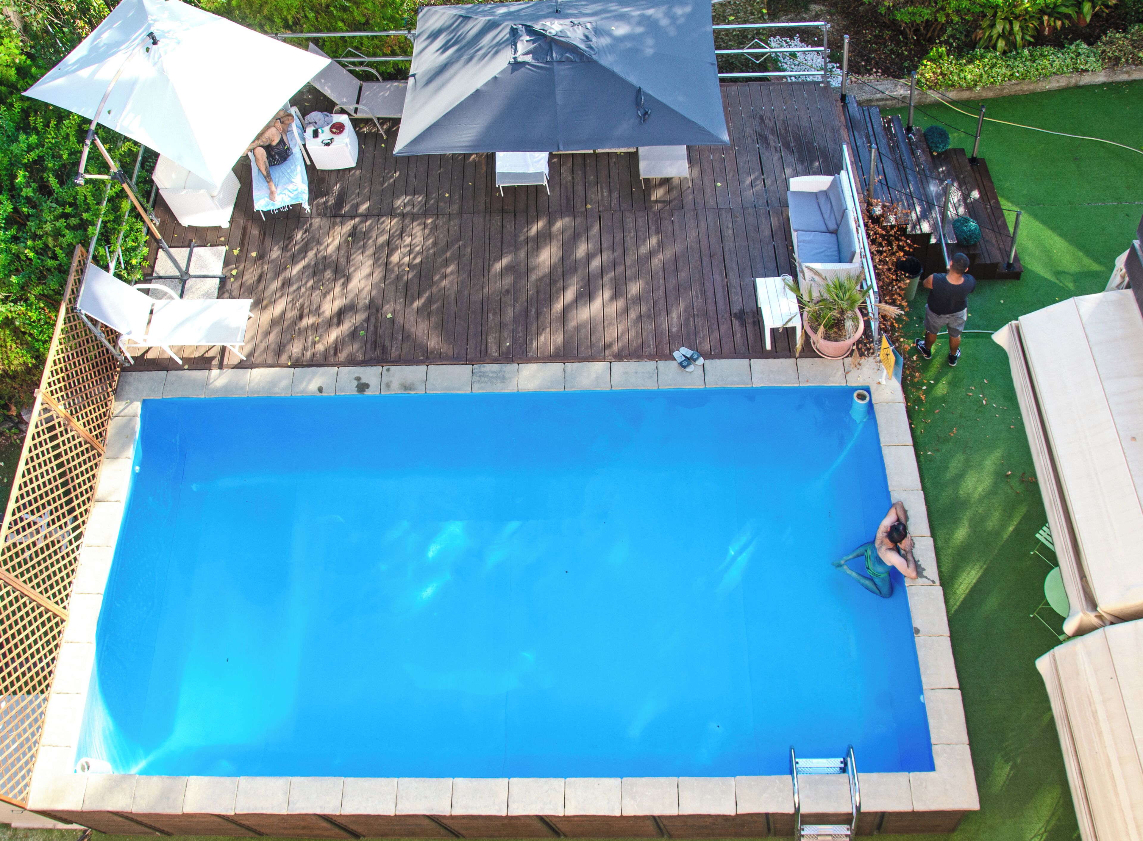 A rectangular swimming pool with a wooden deck surrounding it, equipped with outdoor furniture and umbrellas.