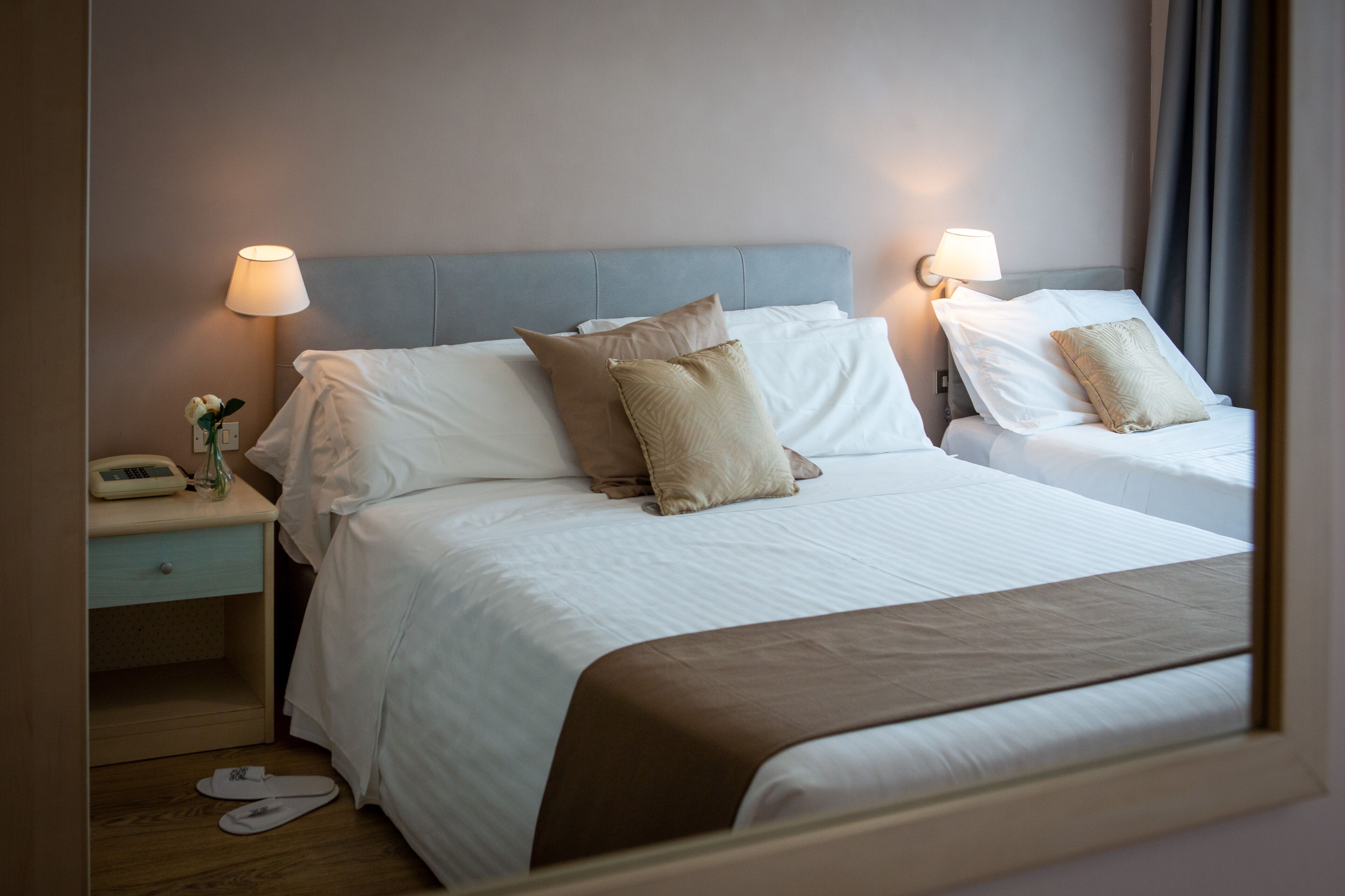 Quarto de hotel com duas camas, cada uma com roupa de cama branca e almofadas bege. Há luminárias de cabeceira, mesas de cabeceira com telefone e um par de chinelos no chão.