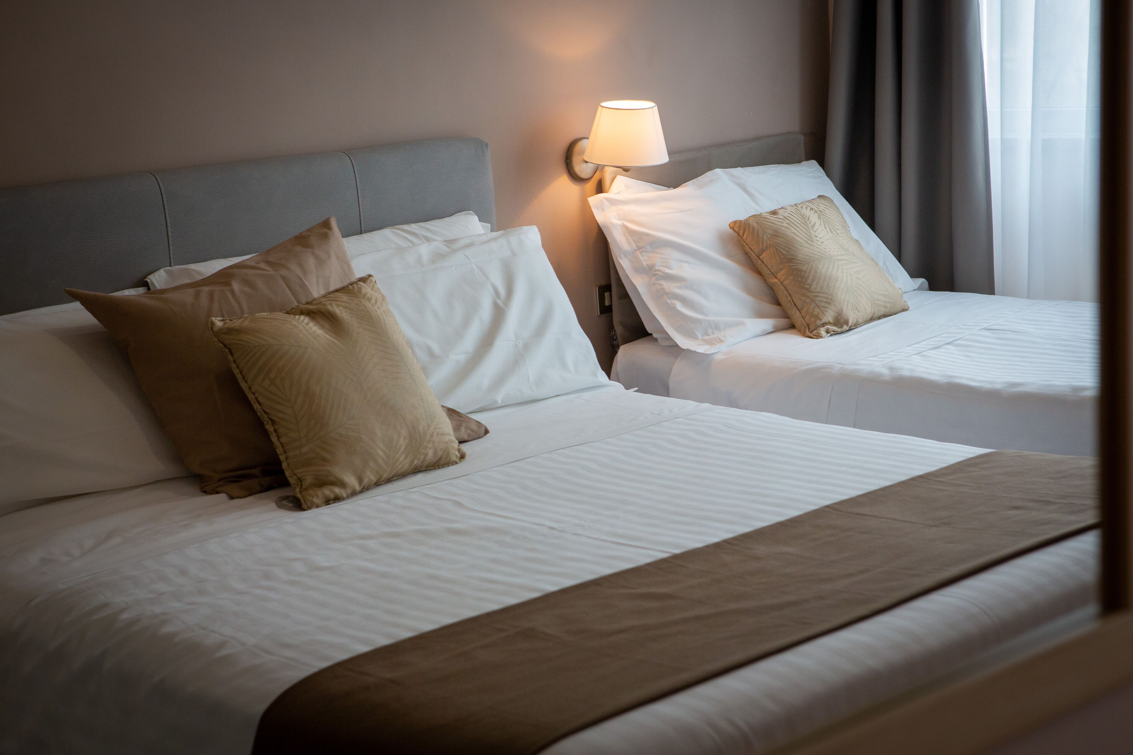 Une chambre d’hôtel avec deux lits, chacun étant équipé de draps blancs et d’oreillers beiges. Une lampe fixée au mur se trouve du côté gauche des lits.