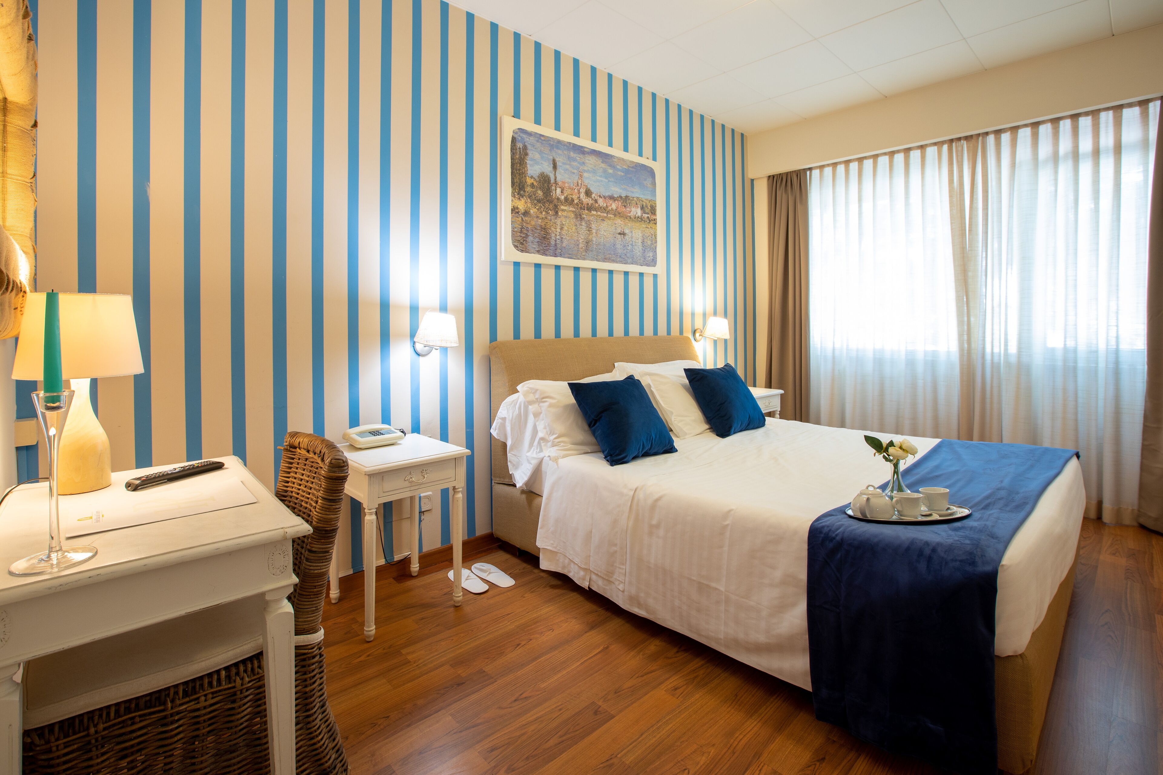 A hotel room with a bed, bedside tables, a nightstand, a lamp, a painting on the wall, and a window with curtains.