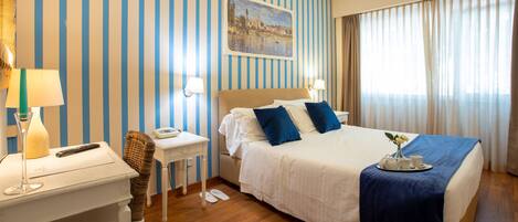A hotel room with a bed, bedside tables, a nightstand, a lamp, a painting on the wall, and a window with curtains.