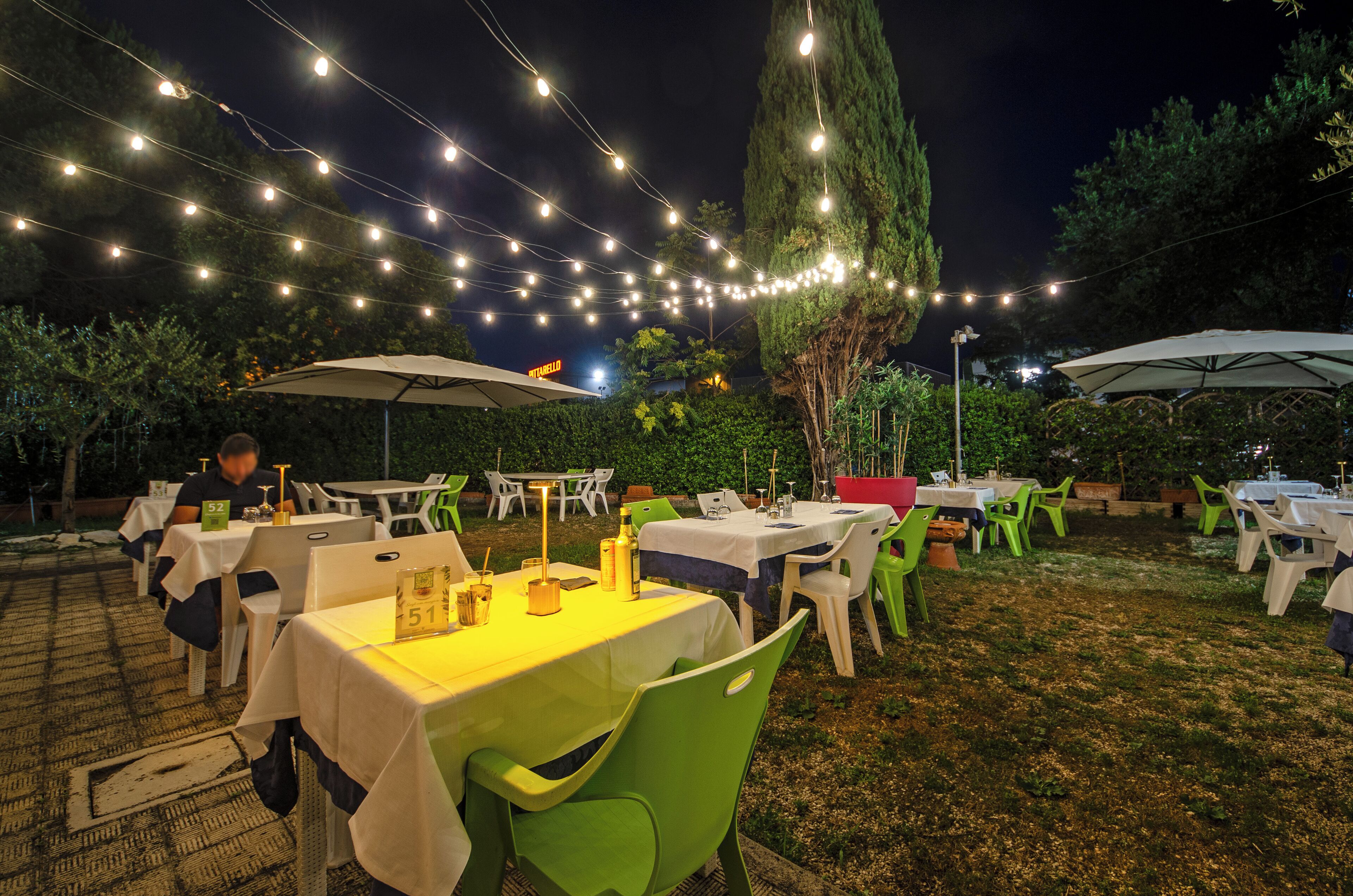 Un espace de restauration en plein air avec des tables et des chaises, des guirlandes lumineuses suspendues et un grand parasol pour faire de l’ombre.