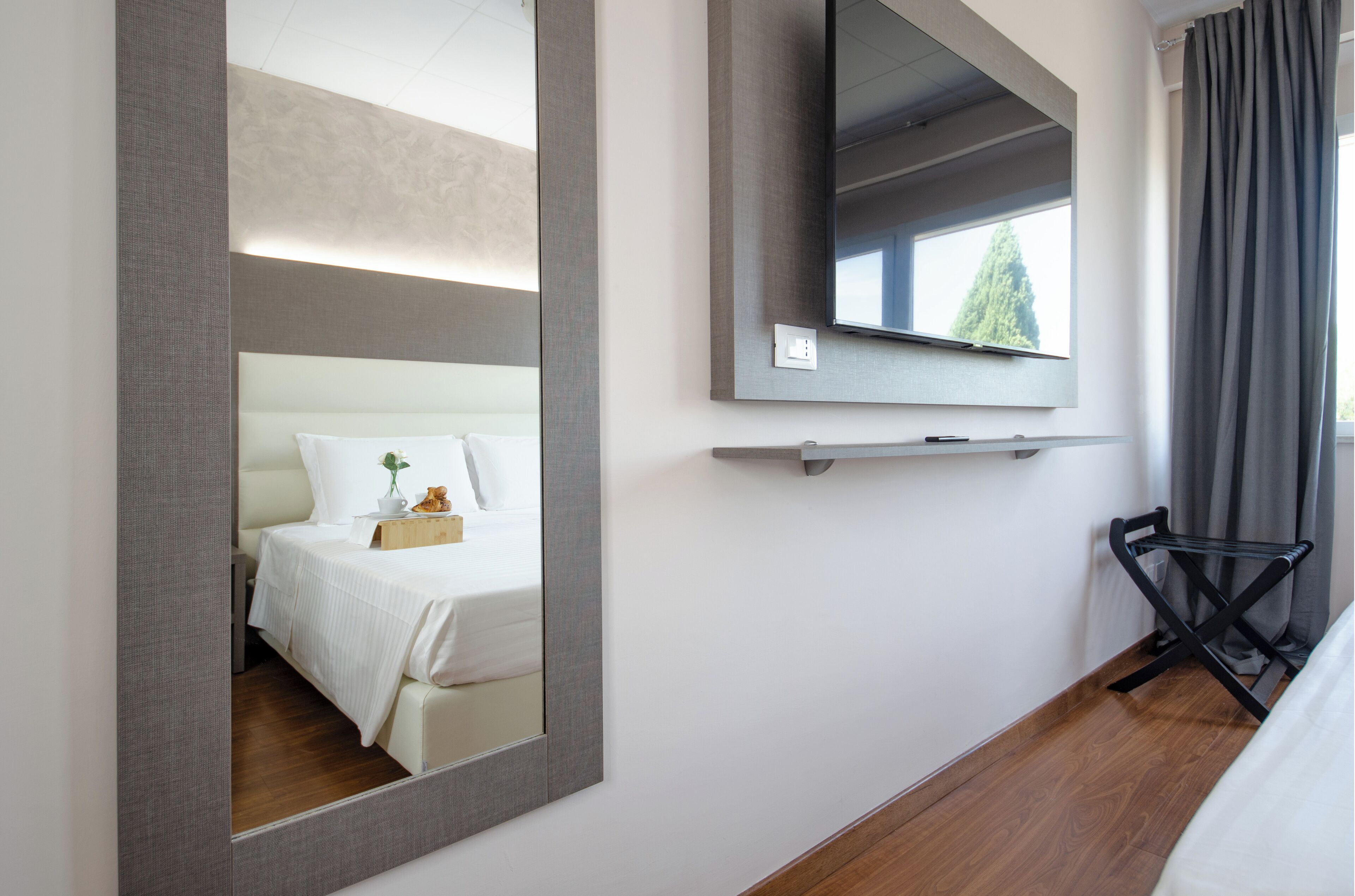Habitación de hotel con un espejo grande, una cama con sábanas blancas, un televisor montado y una mesita de noche con un florero y una planta.