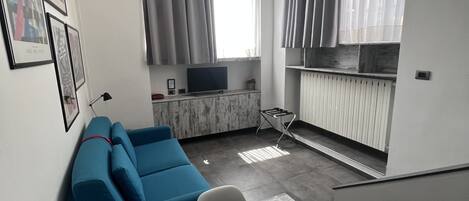 Un salón moderno con un sofá azul, una mesa de comedor con una botella y controles remotos, un televisor y una zona de cocina con un radiador.