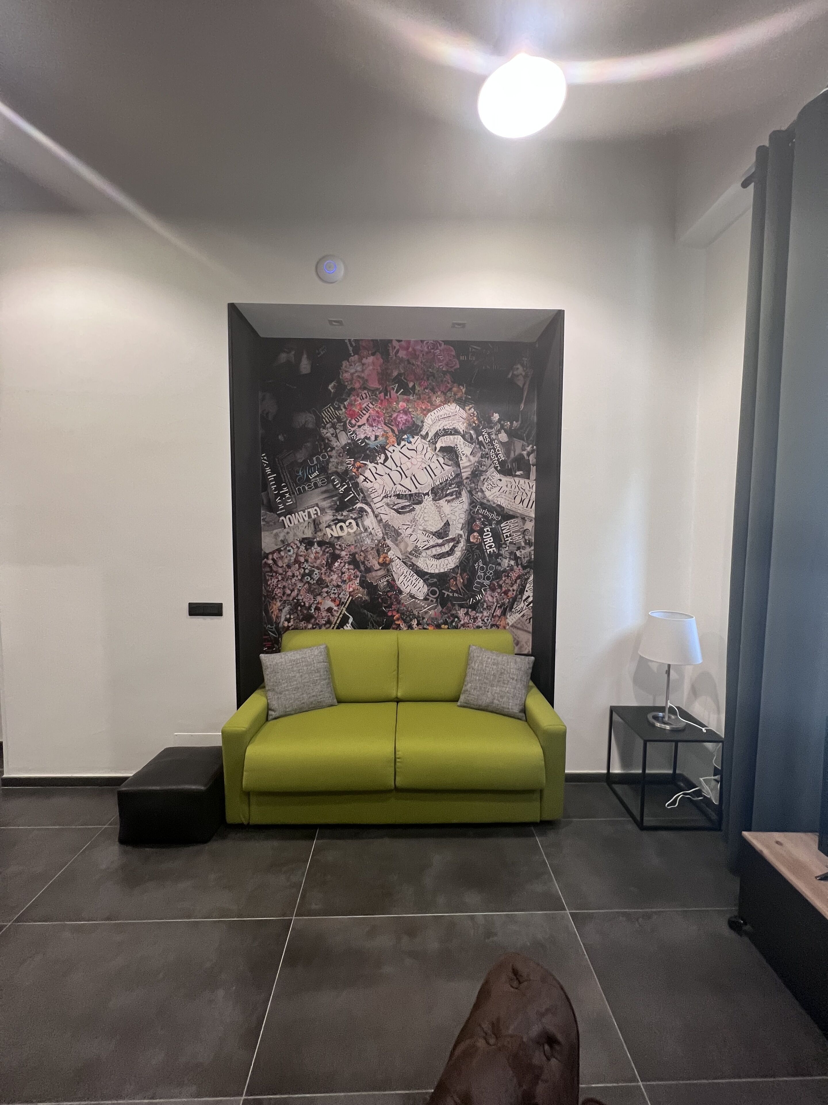 A modern living room with a green sofa, a black ottoman, a small side table with a lamp, and a large abstract wall art piece.