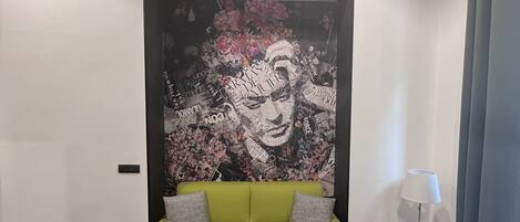 A modern living room with a green sofa, a black ottoman, a small side table with a lamp, and a large abstract wall art piece.