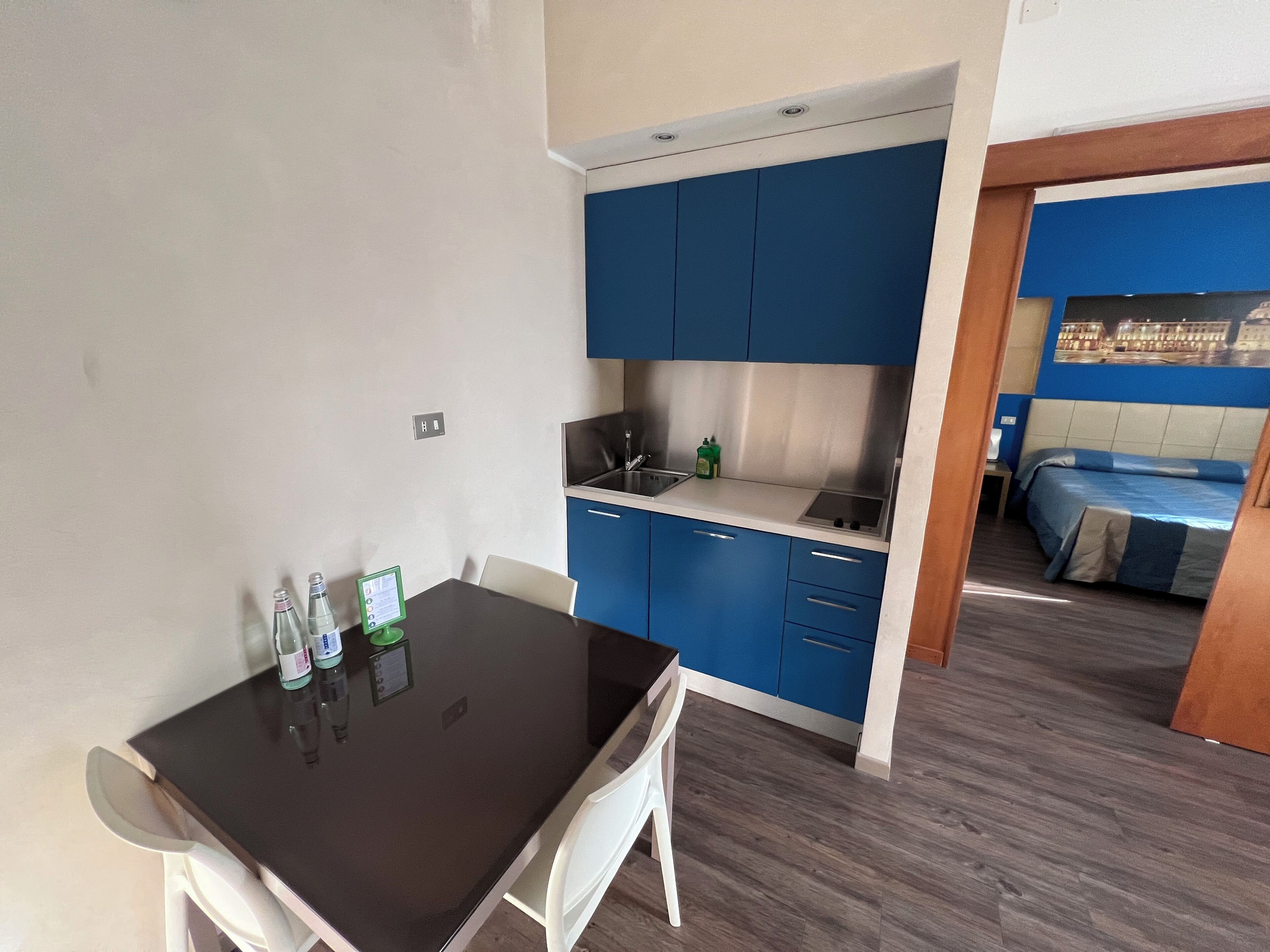 A compact room with a kitchen area featuring blue cabinets and a modern sink. There is a dining table with chairs, and a bed with a blue headboard is visible in the adjacent room.
