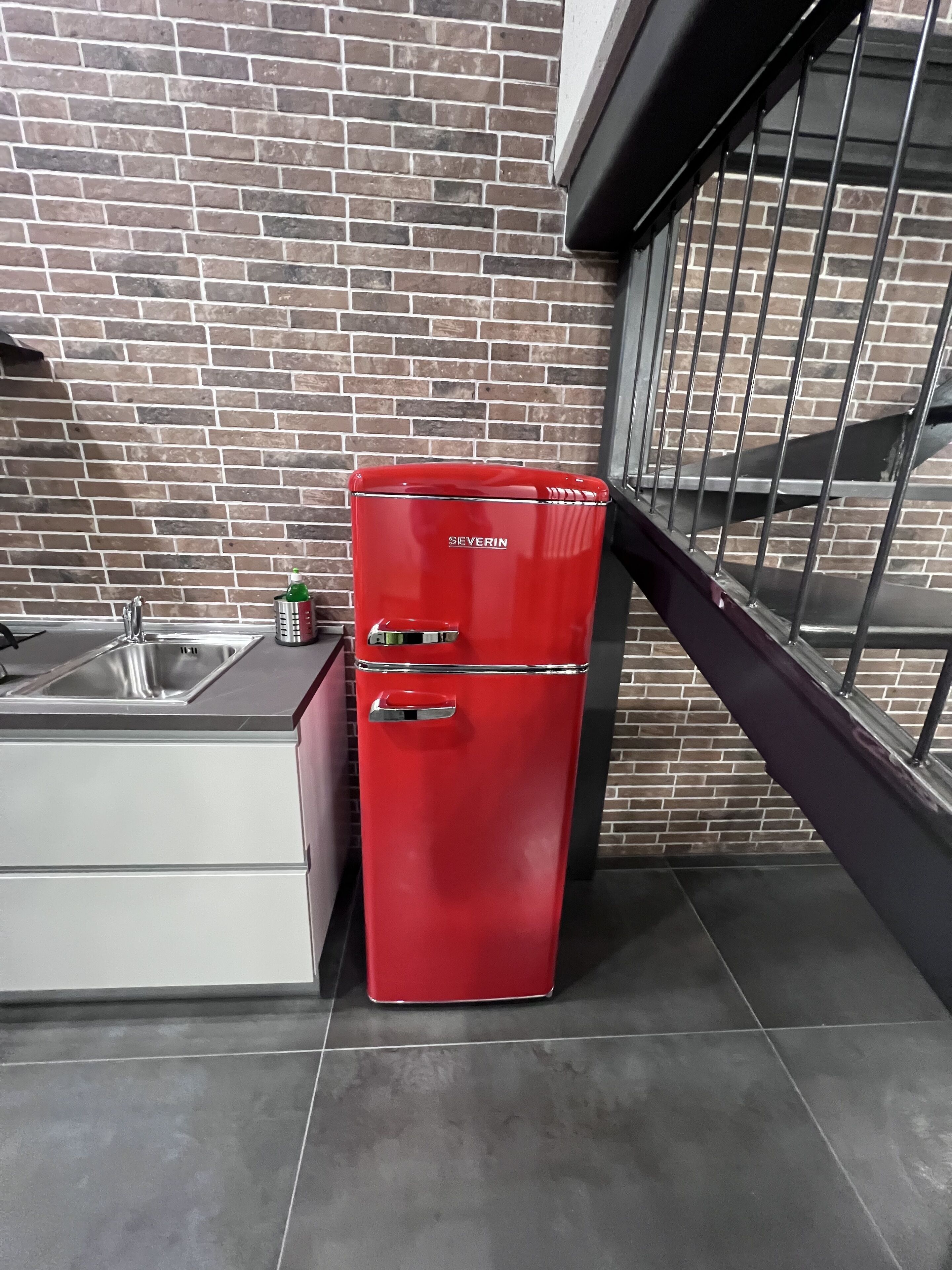 Ein roter Kühlschrank in einer modernen Küche mit Backsteinwand und einer Metalltreppe.