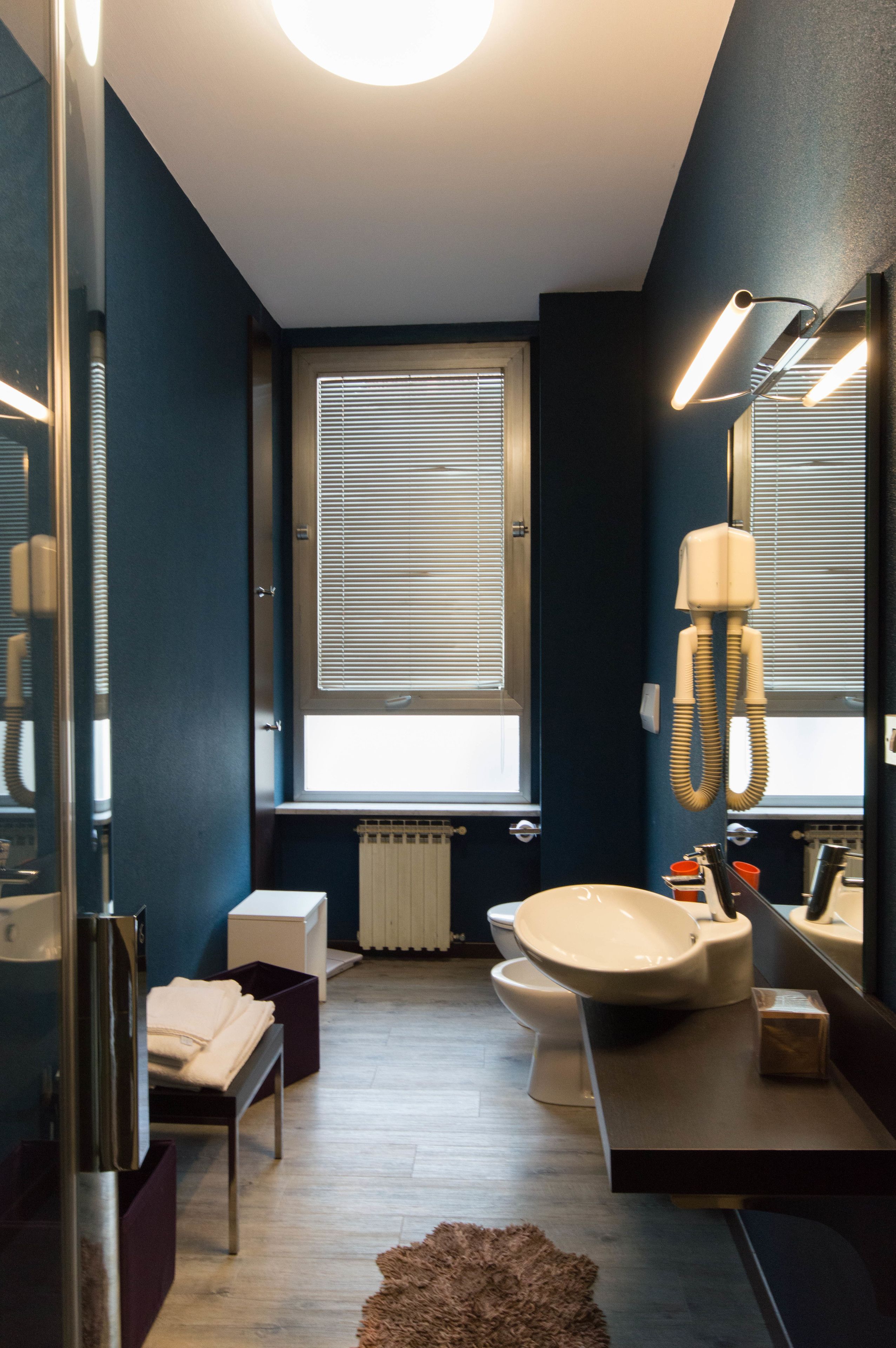 A modern bathroom with a toilet, sink, and mirror. There is a window with blinds, a radiator, and a hairdryer on the wall.