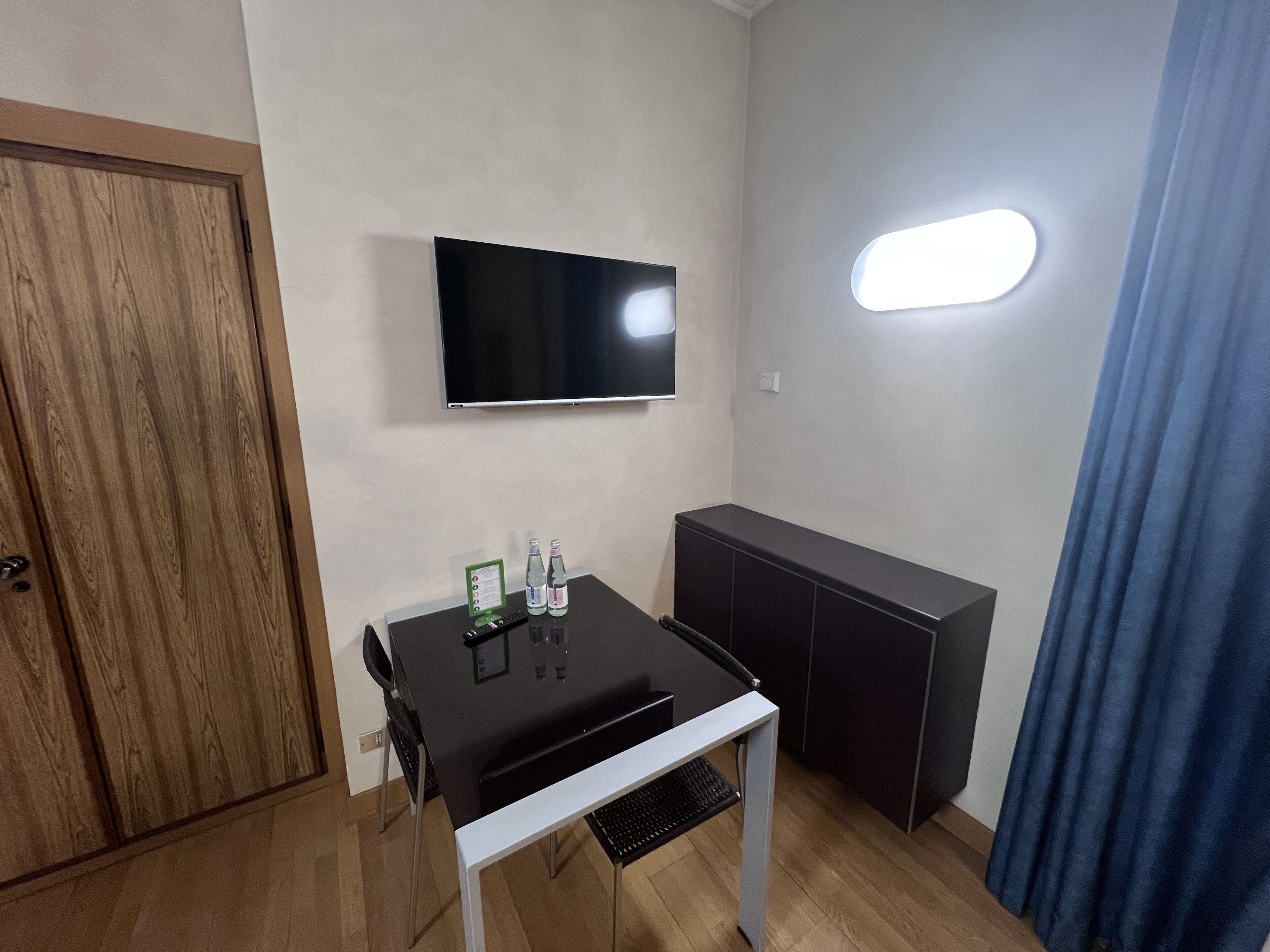 A small room with a wooden door, a flat-screen TV mounted on the wall, a small table with a remote control and bottles, and a dark cabinet.