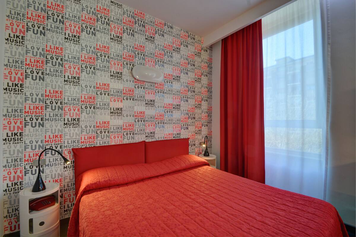 Une chambre d’hôtel avec une couvre-lit rouge, un mur sur lequel on peut lire « J’aime la musique », une table de chevet avec une lampe et une fenêtre avec des rideaux rouges.