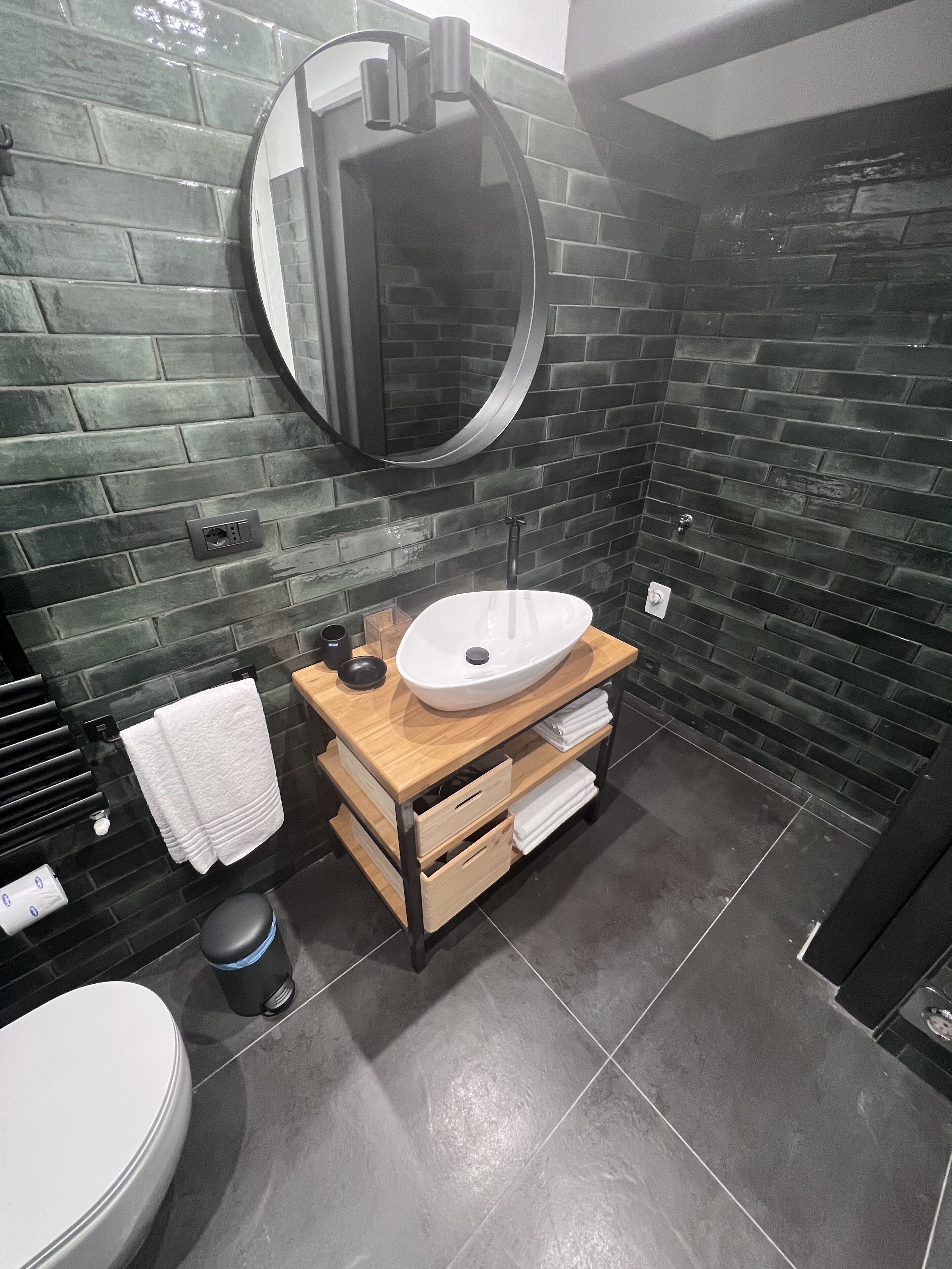 Een moderne badkamer met een muur van zwarte bakstenen, een houten wastafelmeubel met een witte wasbak, een ronde spiegel en een toilet.