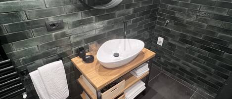Un baño moderno con pared de ladrillo negro, tocador de madera con lavamanos blanco, espejo redondo y un inodoro.