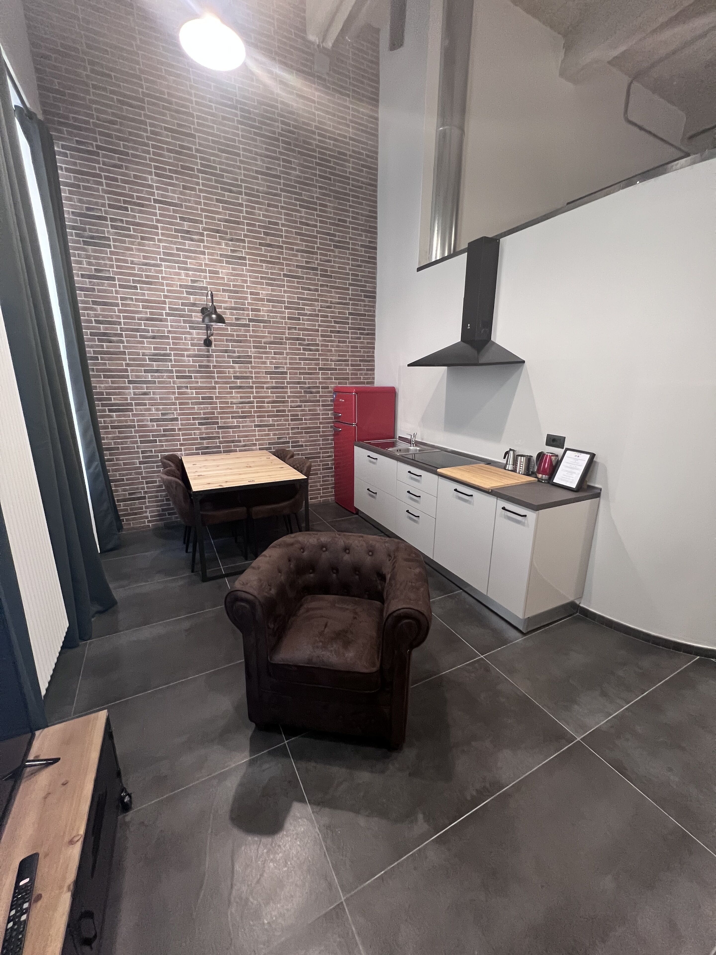 A compact kitchen with a brick wall, a red refrigerator, a dining table with chairs, and a tufted armchair.