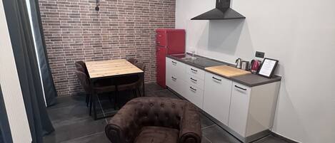 A compact kitchen with a brick wall, a red refrigerator, a dining table with chairs, and a tufted armchair.