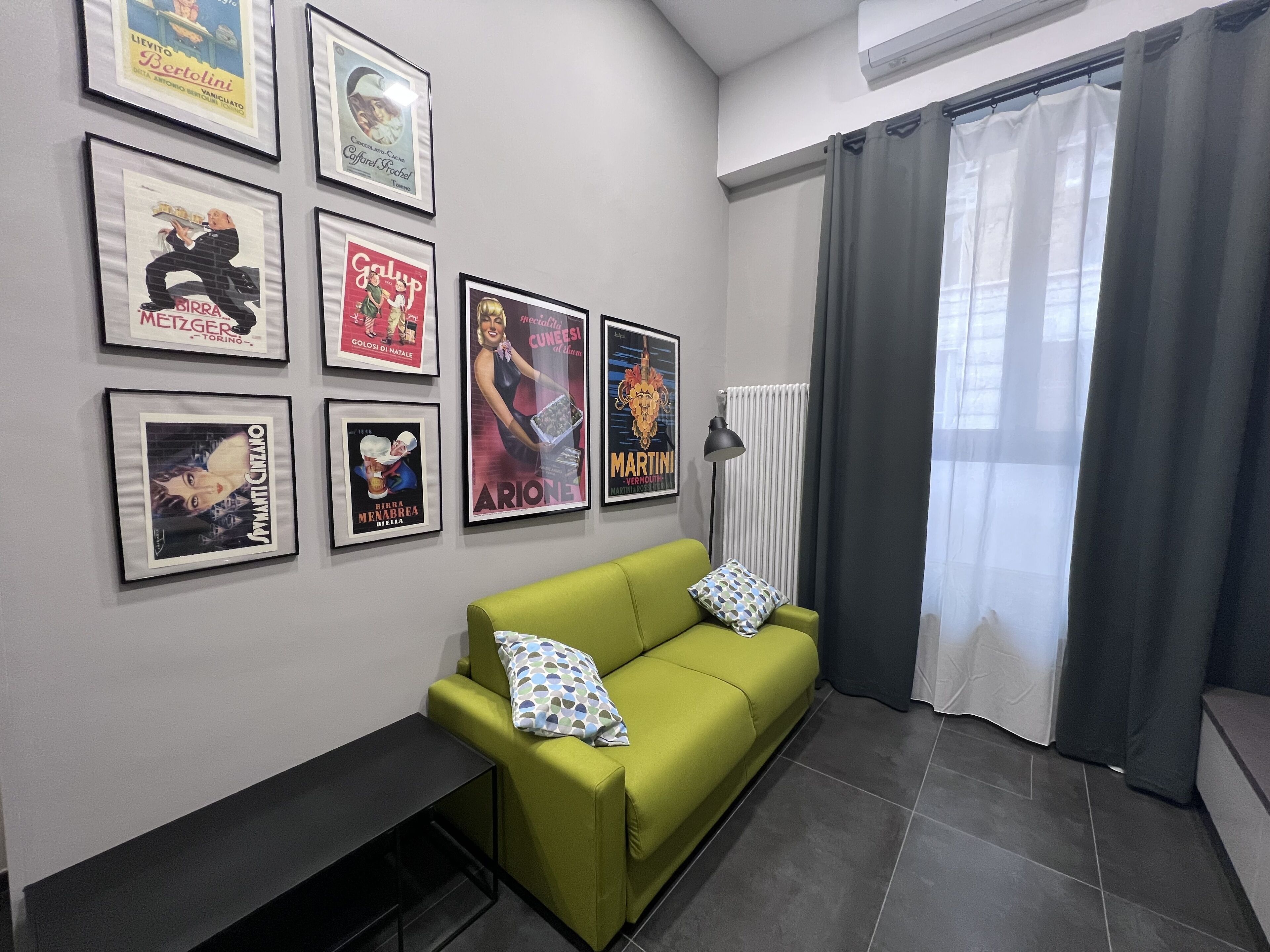 A living room with a green sofa, a black side table, and a radiator. There are framed posters on the wall.