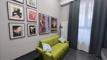 A living room with a green sofa, a black side table, and a radiator. There are framed posters on the wall.