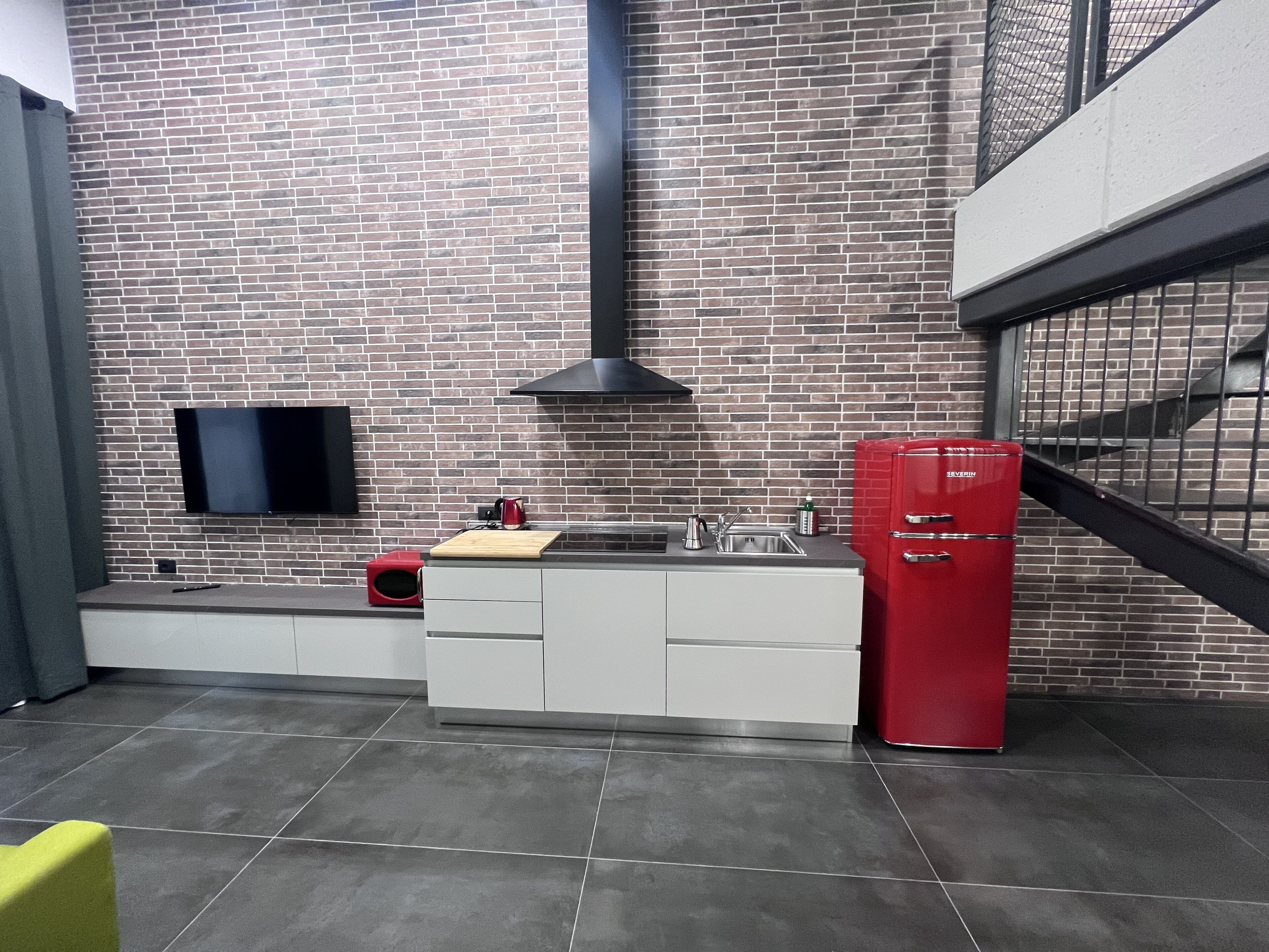 Una cocina moderna con protector mural de ladrillo, un televisor de pantalla plana, un refrigerador rojo y una tabla de cortar de madera sobre la encimera.