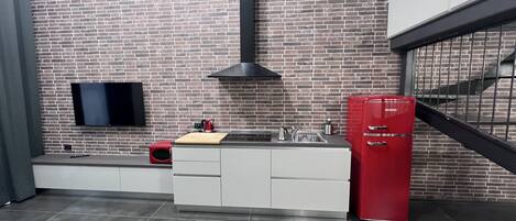 Una cocina moderna con protector mural de ladrillo, un televisor de pantalla plana, un refrigerador rojo y una tabla de cortar de madera sobre la encimera.