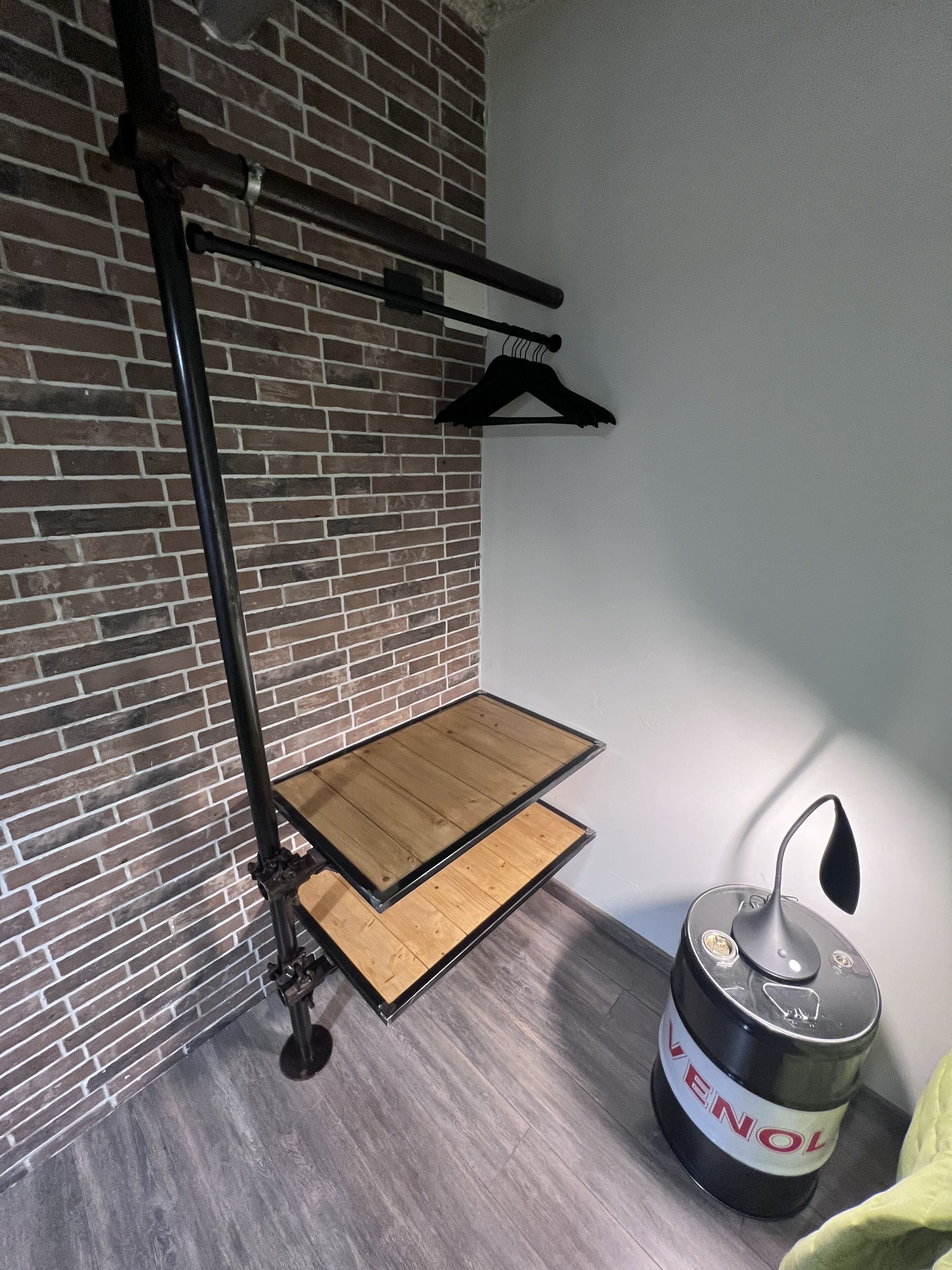 Een kamer met een bakstenen muur, een houten platform, een metalen rek, een zwarte prullenbak en een groene deken.