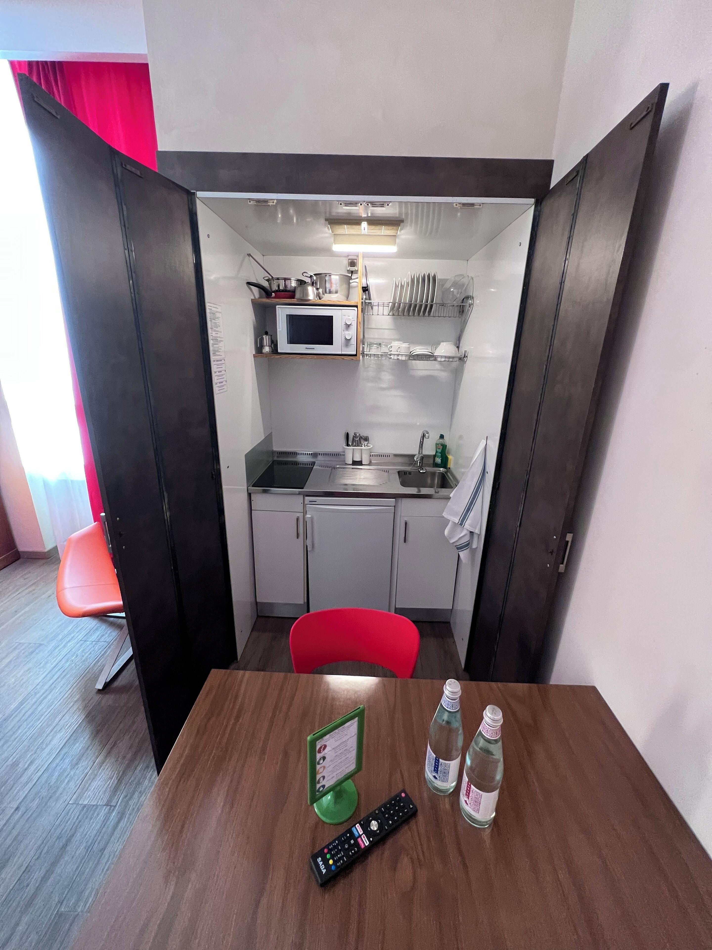 Une petite cuisine équipée d’un micro-ondes, d’un évier et de placards de rangement. Une chaise rouge est visible au premier plan, et une télécommande se trouve sur la table.