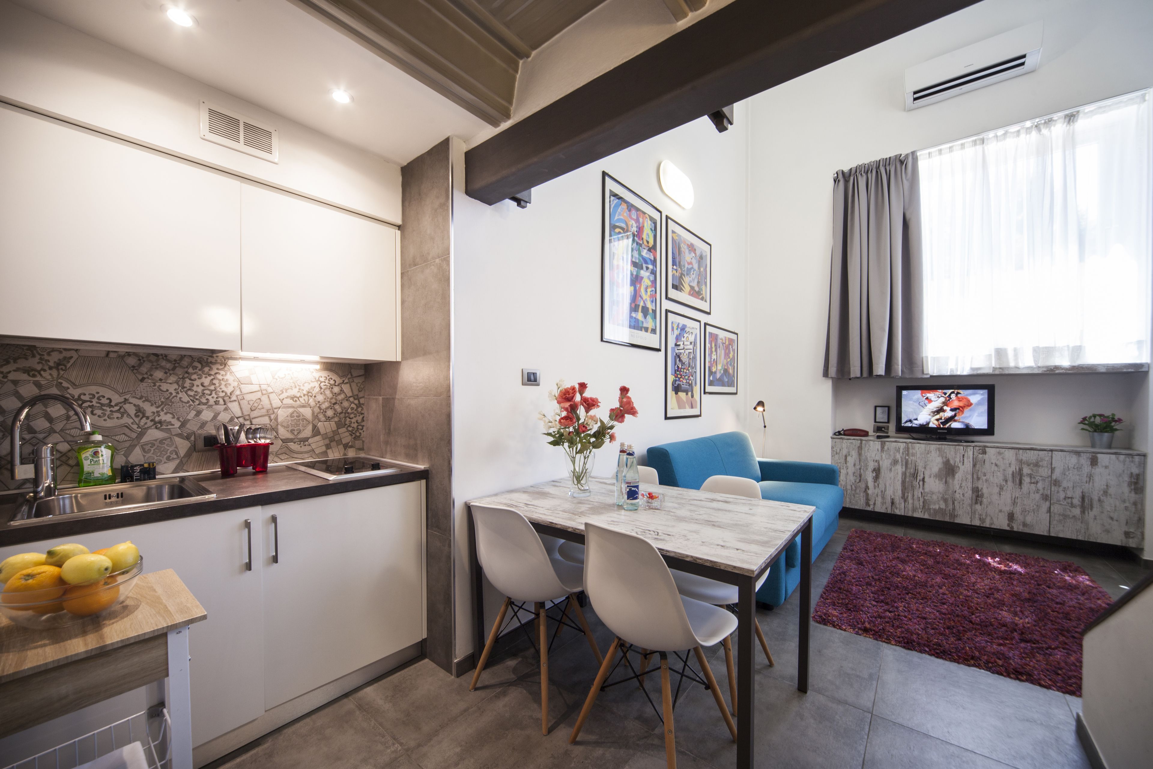 Una cocina moderna con armarios blancos, encimera de mármol y un comedor con mesa y sillas. Hay un televisor sobre un mueble de madera, y una alfombra roja en el suelo.