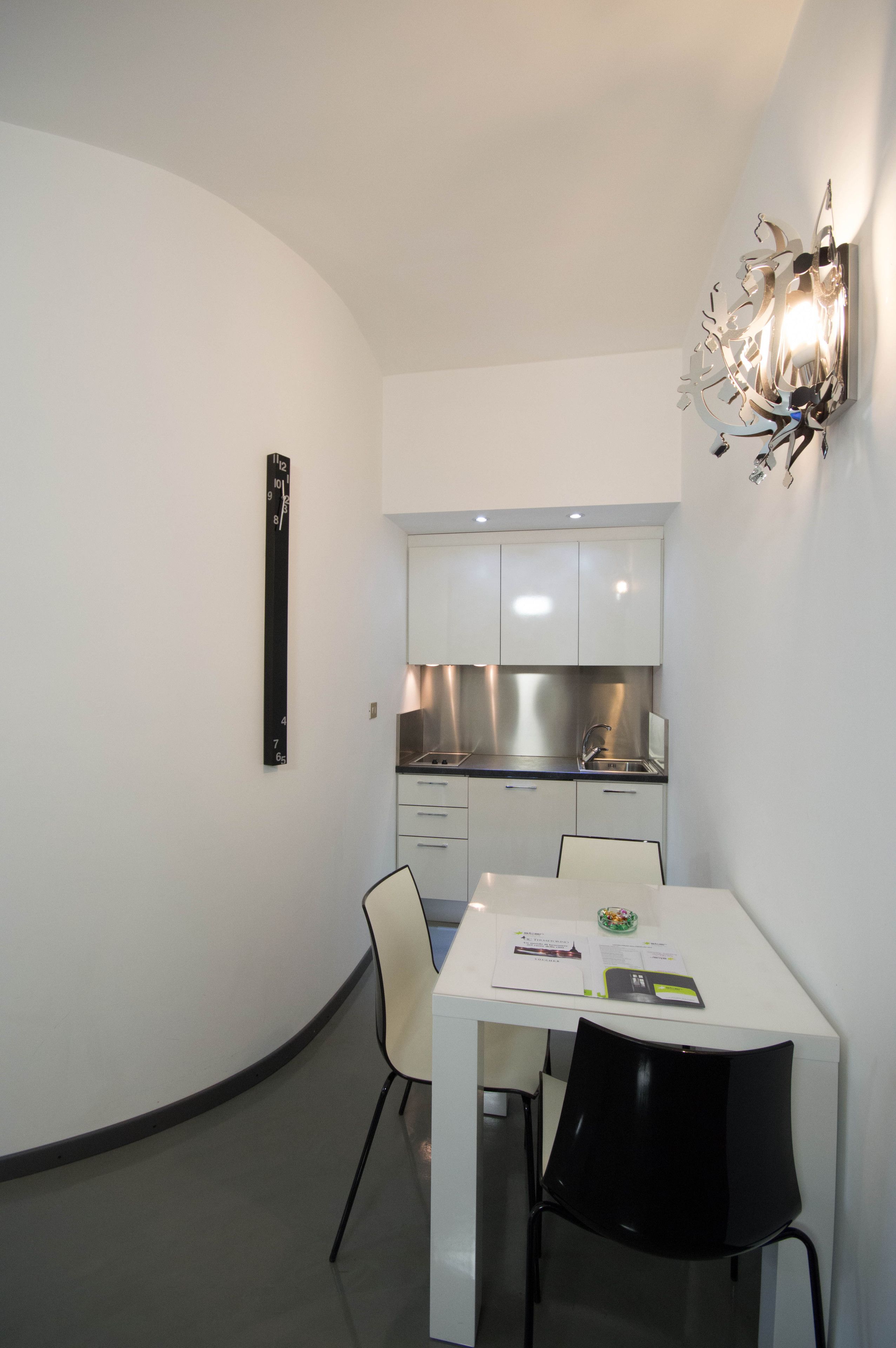 A compact kitchen with white cabinets and stainless steel appliances, a white dining table with two chairs, and a wall-mounted light fixture.