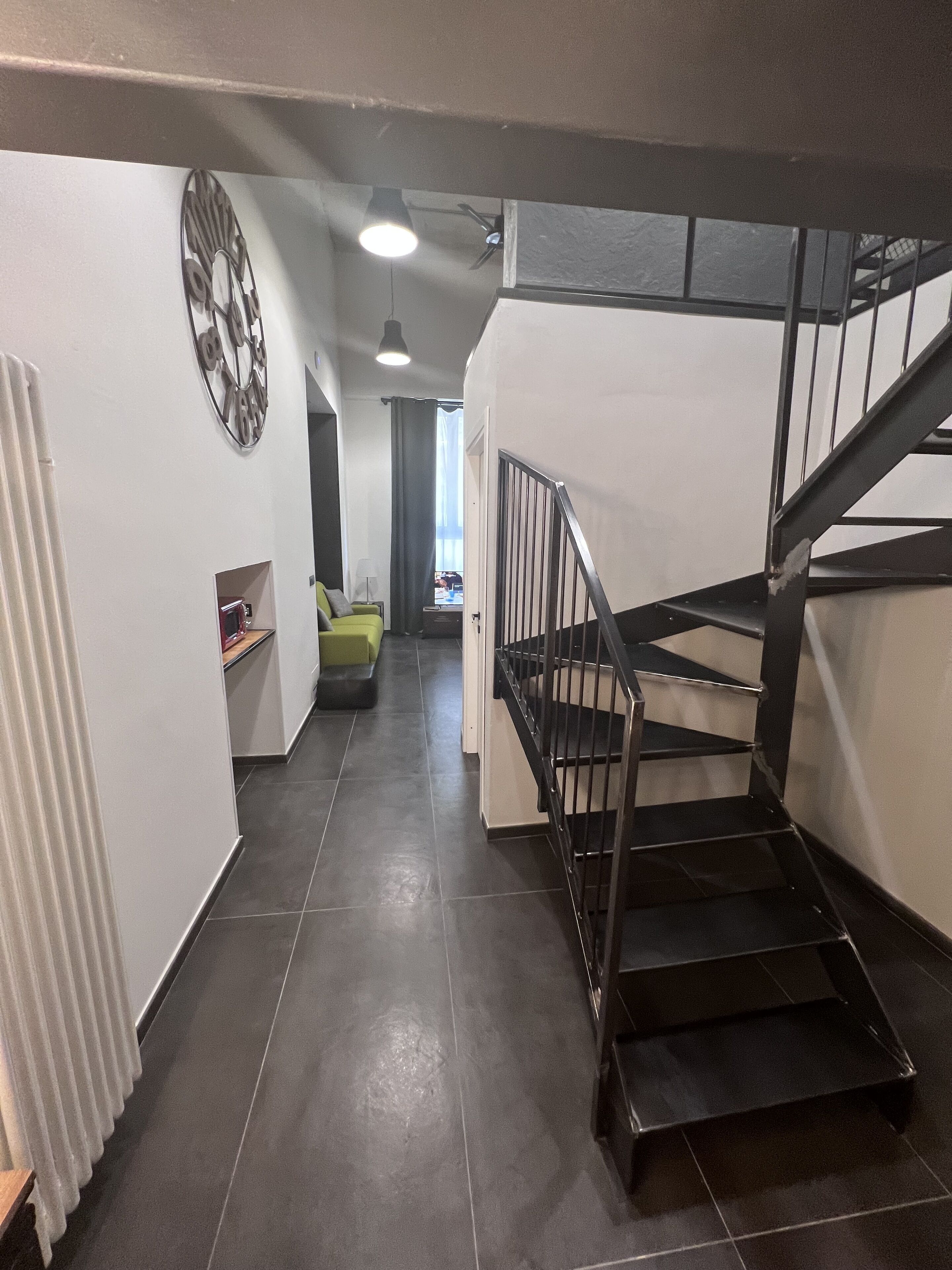 A staircase with a metal railing leading to an upper floor. A radiator is visible on the left, and a door opens to a room with a desk and chair.