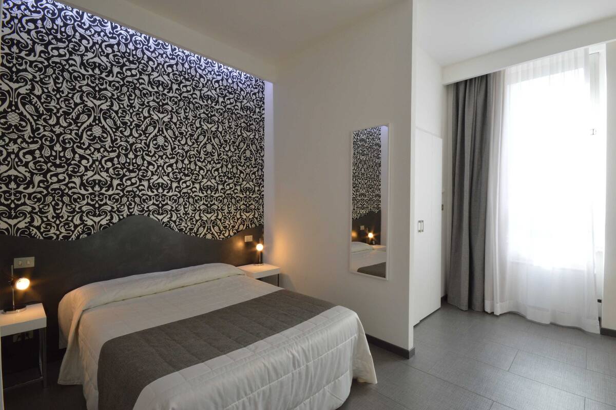 Une chambre d’hôtel avec un grand lit, des tables de chevet avec des lampes, un mur décoré d’un motif, un miroir et une grande fenêtre avec des rideaux.