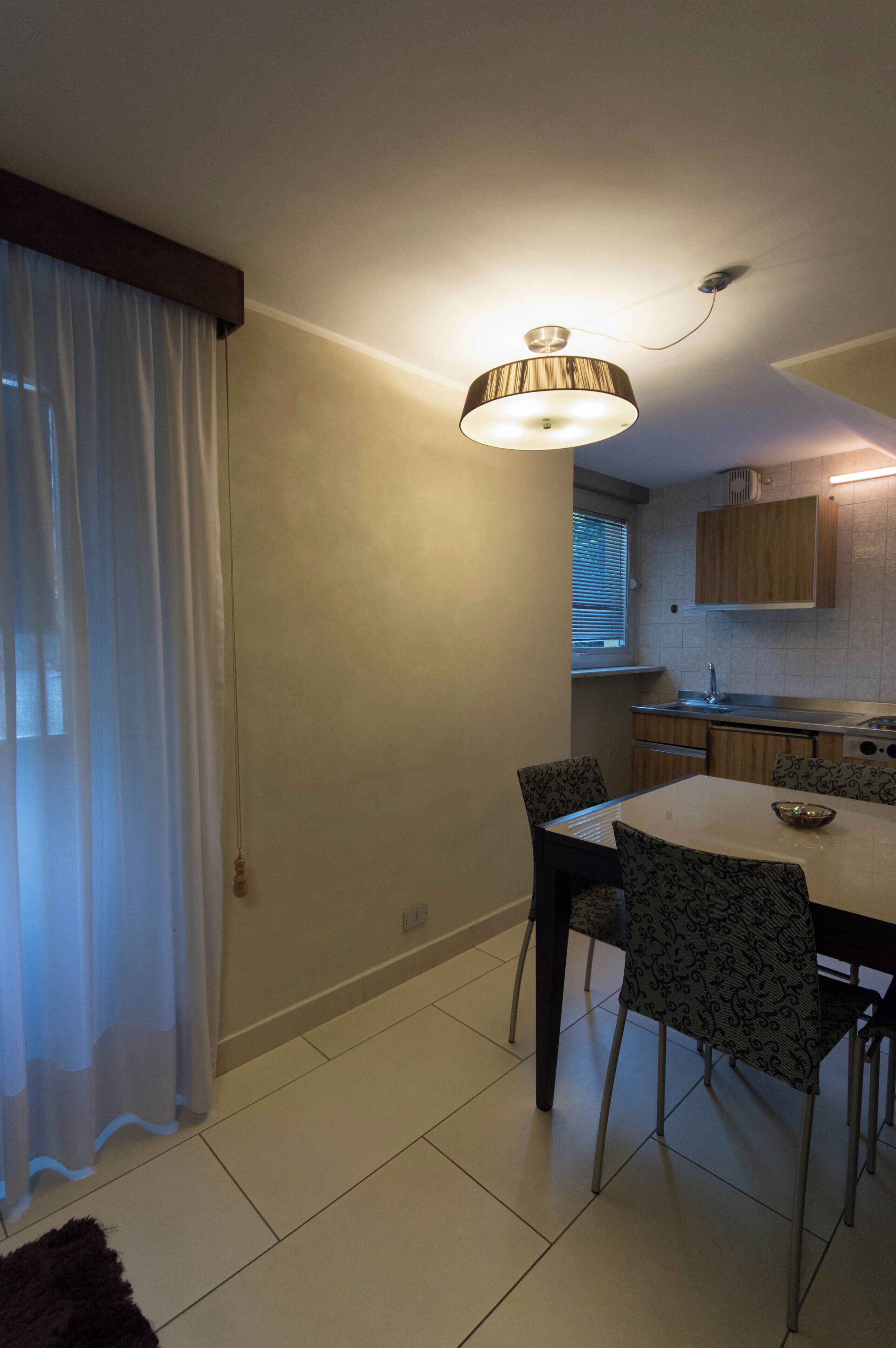 Un comedor con una mesa y sillas, un lámpara colgante, una ventana con cortinas y una cocina al fondo.