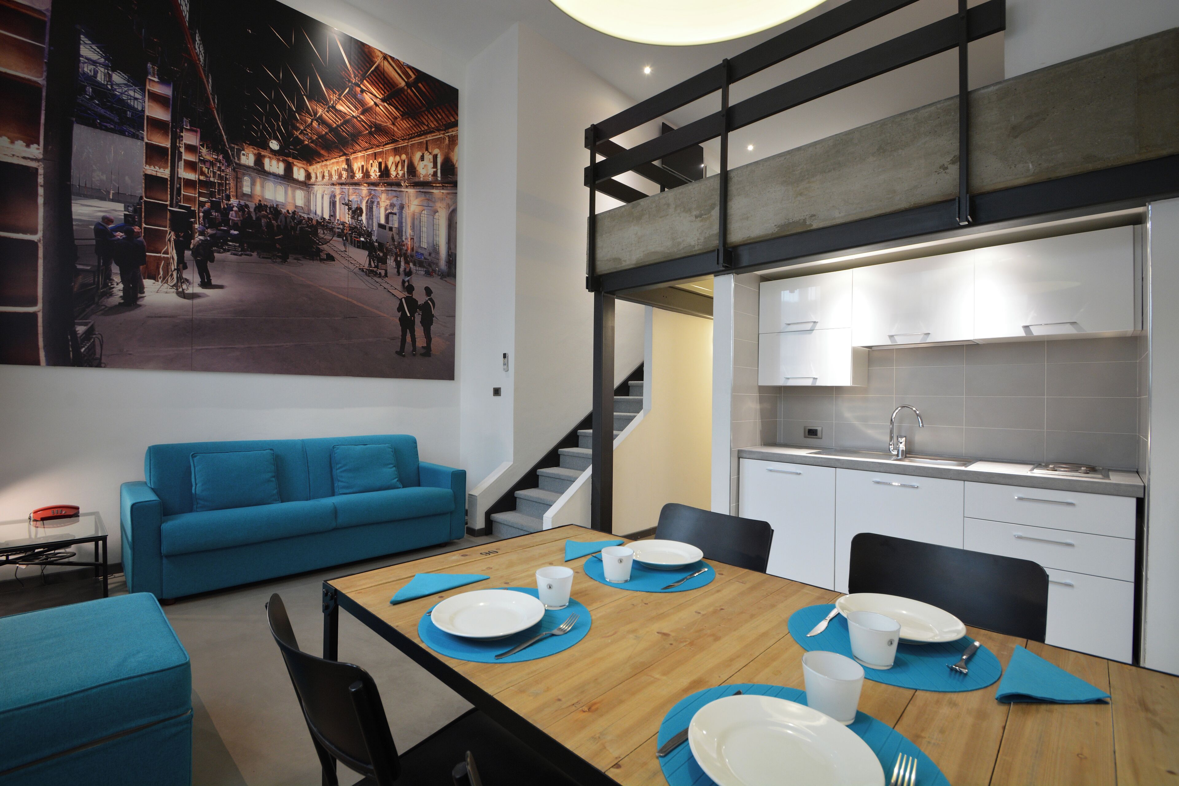 A modern dining area with a wooden table set for four, blue placemats, and white plates. A blue sofa and black chairs are in the background.