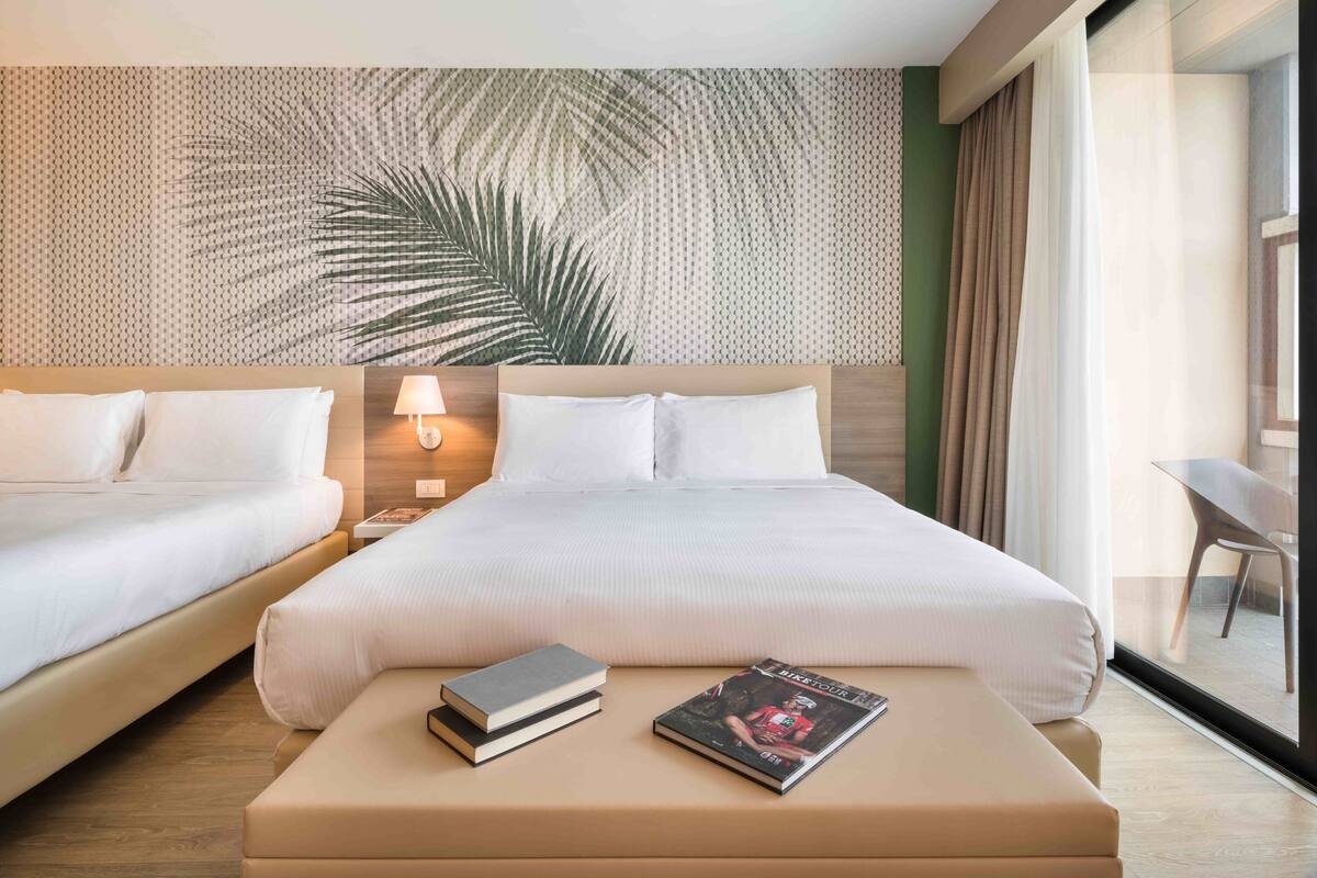 A hotel room with two beds, a nightstand with a lamp, a coffee table with books, and a large window with a view.
