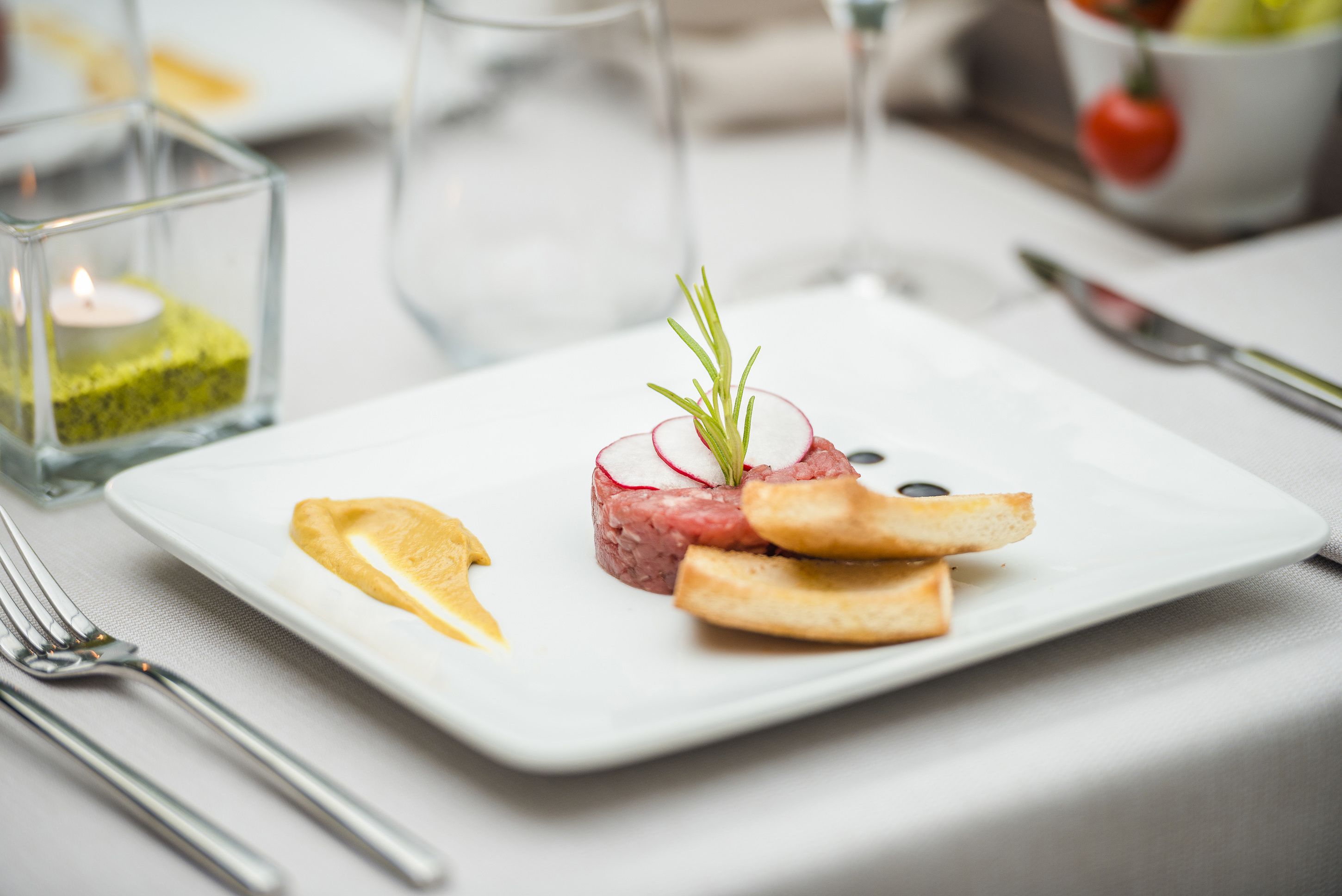 A white plate with a dish of steak tartare, lemon slices, and a sprig of rosemary.
