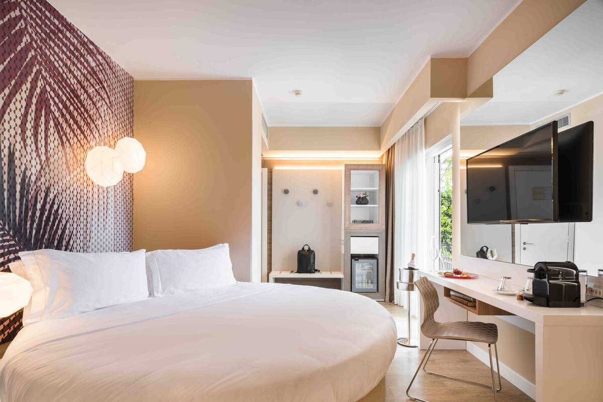 A hotel room with a large bed, a desk with a flat-screen TV, a minibar, and a seating area.