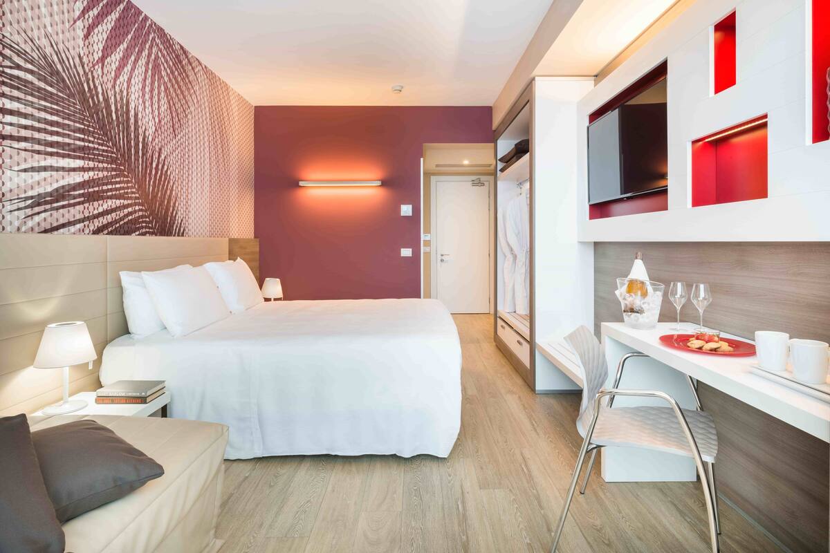 A modern hotel room with a bed, bedside table, lamp, and a small seating area. There is a kitchenette with a microwave, toaster, and a glass of water. The room has a large window and a decorative wall panel.