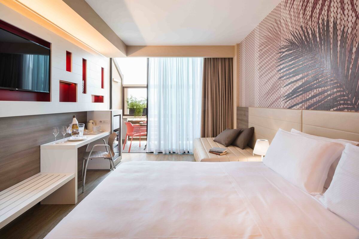 A modern hotel room with a large bed, a desk with a chair, a TV, and a view of the outdoors.
