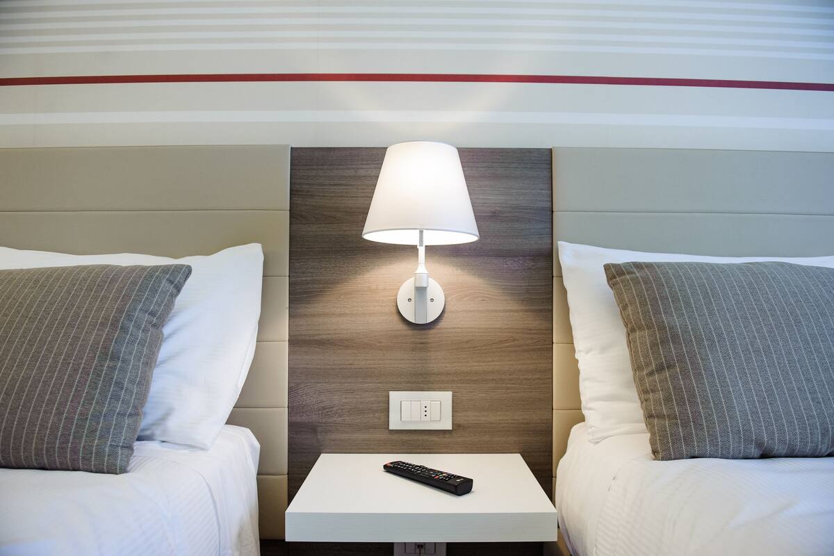 A hotel room with two beds, a nightstand with a remote control, a wall-mounted lamp, and a power outlet.